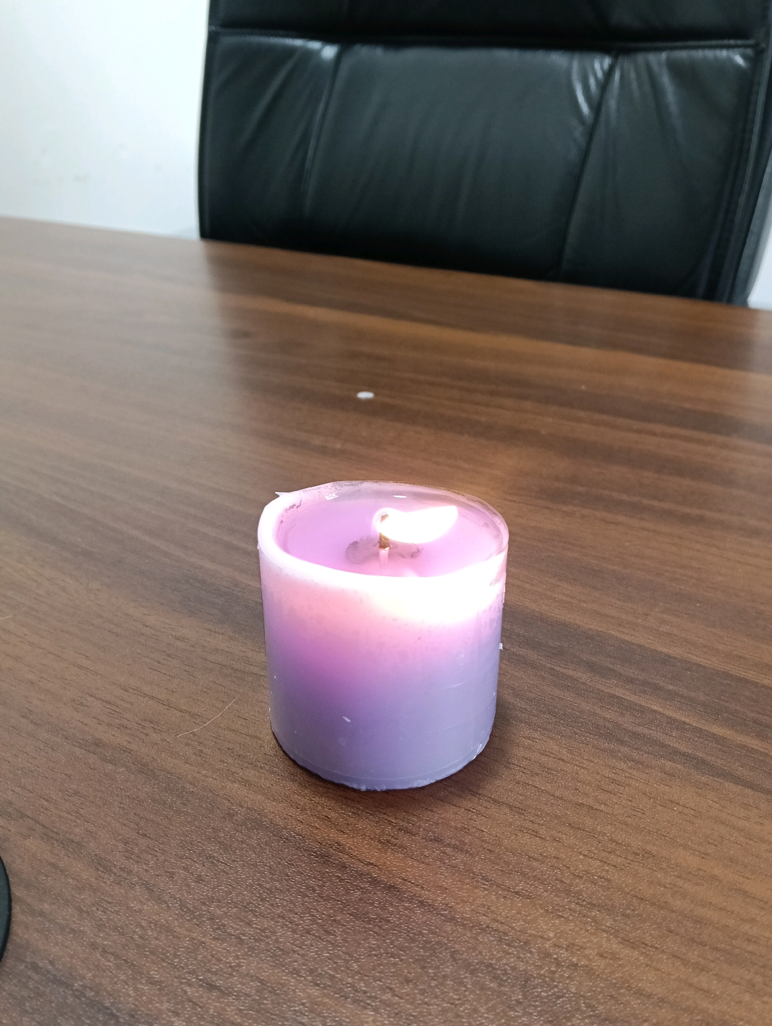 A lit, cylindrical, lavender-colored candle rests on a dark wood table. The candle's flame casts a warm glow. In the background, the smooth, black leather of an office chair is visible, with a portion of a white wall to the left. This scene is located within an office environment in Nairobi, Kenya.