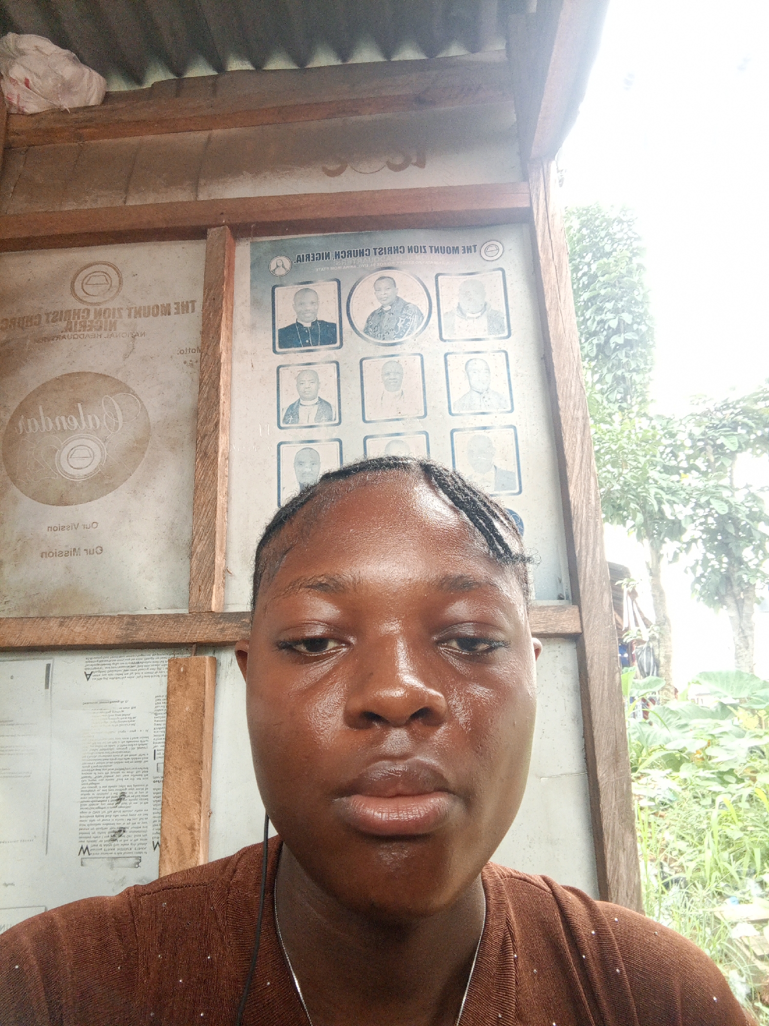 A close-up of a woman with a brown shirt, facing the camera. She appears to be standing next to a wooden structure with posters and text. One of the signs reads "THE MOUNT ZION CHRIST CHURCH NIGERIA." The surrounding environment shows lush greenery, possibly indicating an outdoor or rural setting. The exact location is Mbribit, Nigeria.