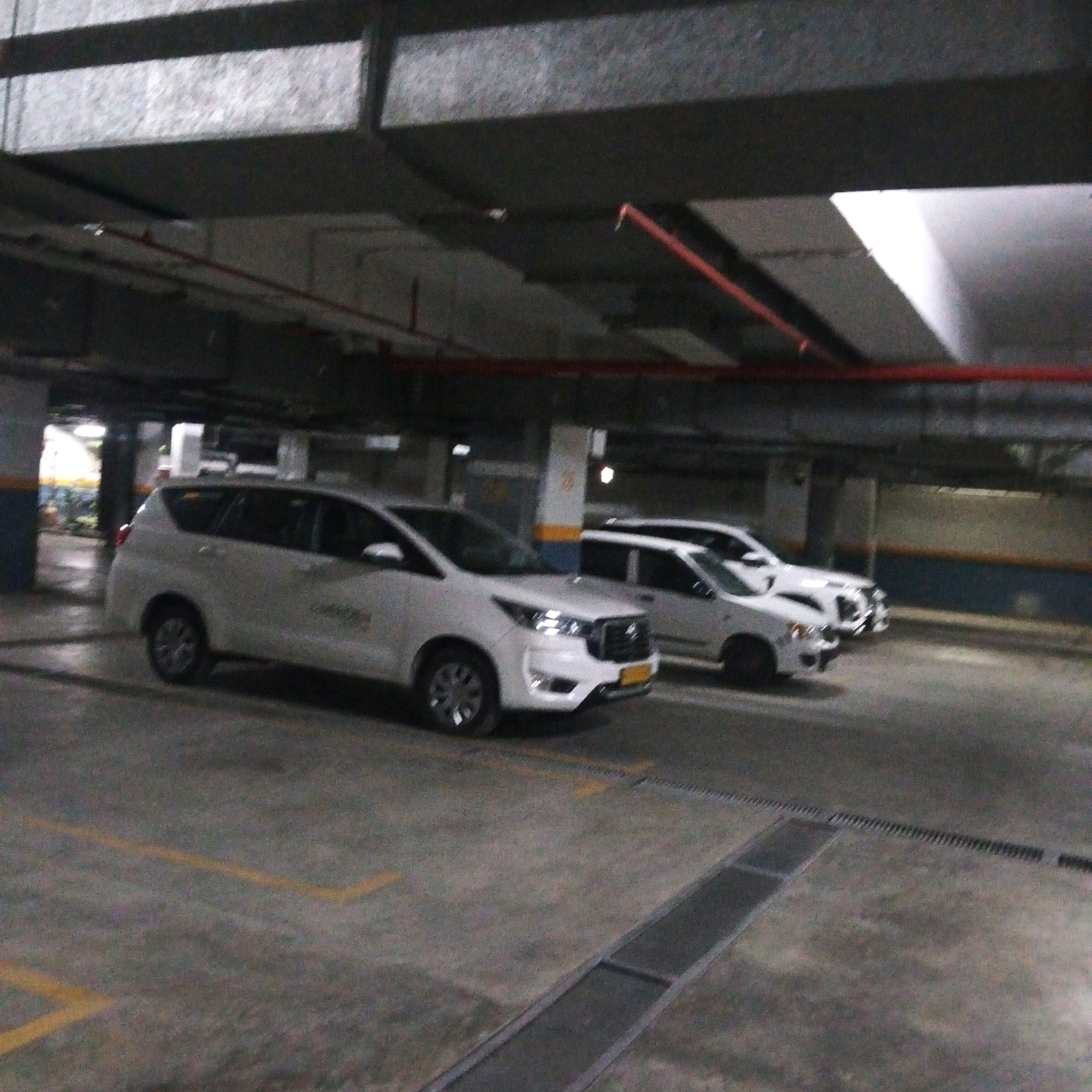 The image captures an indoor parking garage, likely in Bal Sehchandar, India, showing several white vehicles parked in designated spots. The concrete floor is marked with yellow lines, and a metal grate runs along the ground in the foreground. Above, exposed pipes and structural beams are visible. The lighting is dim, typical of an underground parking facility. The primary focus is on the vehicles and the environment of the parking structure.