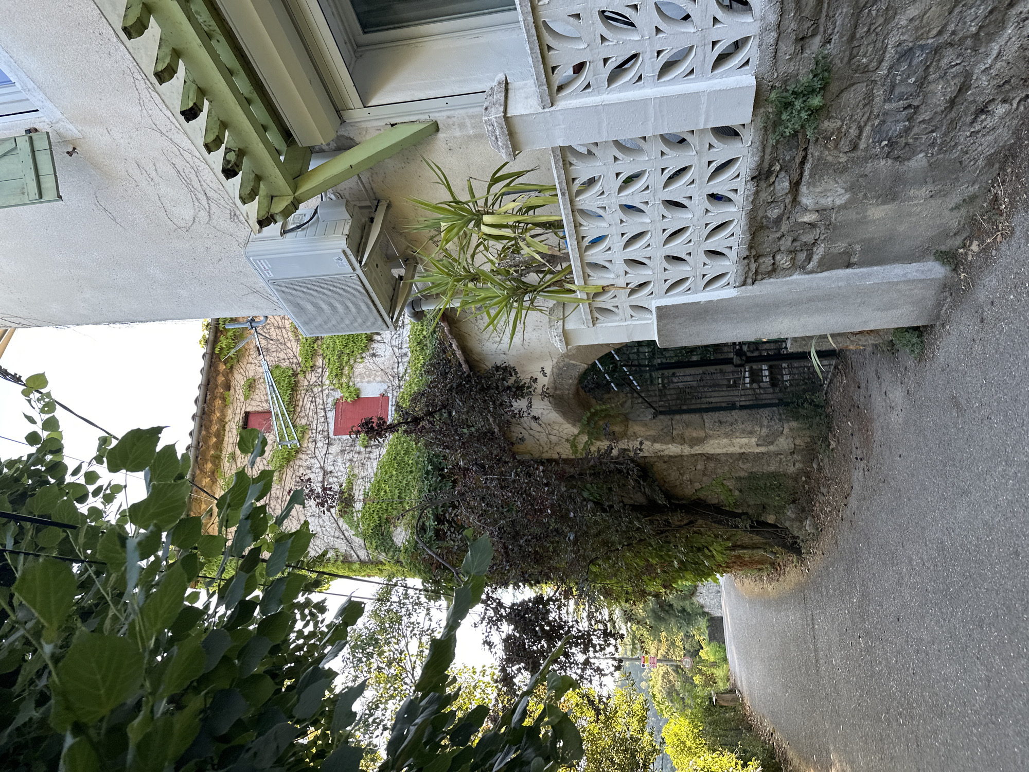 A narrow paved road curves uphill to the left, bordered by a stone wall and lush vegetation. On the right, a stone wall transitions to a lighter stucco wall adorned with ivy and an air conditioning unit. Further along the right, a decorative white concrete balustrade runs parallel to the road. A wrought iron gate is visible within an arched stone entryway in the background. The scene suggests a residential area in the French countryside, possibly Générargues.