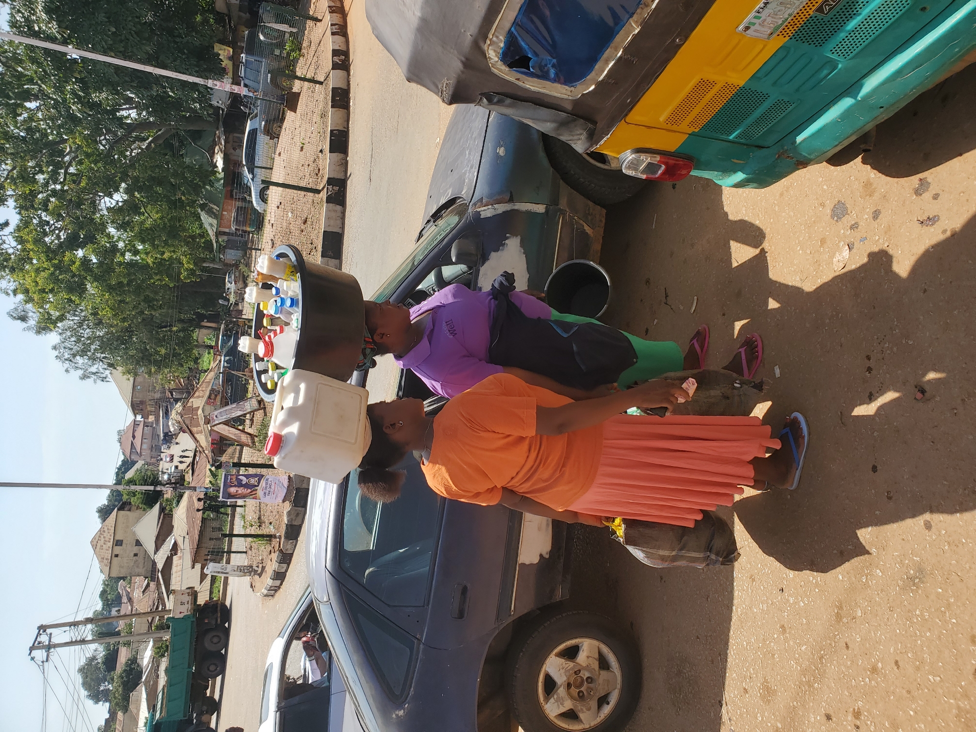 In Bukuru, Nigeria, two women stand beside a dark sedan parked on a dusty street. The woman on the left, wearing an orange t-shirt and a peach skirt, balances a large white plastic container filled with various bottles on her head. She also carries a large dark bag in her right hand. To her right, a woman in a purple polo shirt and green leggings holds a black bucket in her left hand and a black bag in her right. A yellow and teal tricycle is parked to the right of the sedan. In the background, a large green dump truck is visible, along with several buildings, trees, and power lines. The scene appears to be a busy roadside market or thoroughfare.