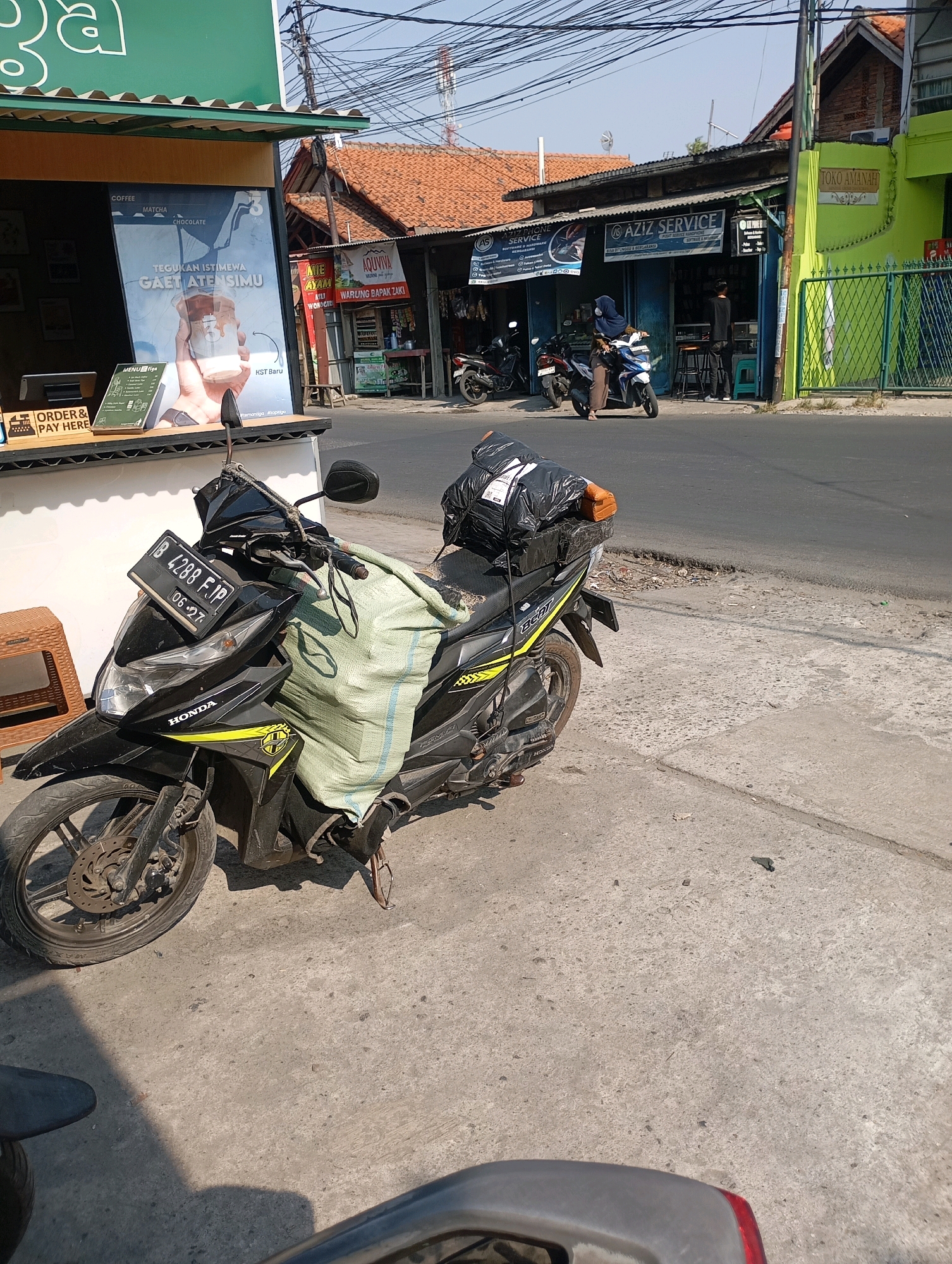 The image captures a street scene in Babelan, Indonesia, under a clear sky. In the foreground, a black Honda motorcycle is parked on a concrete sidewalk. The motorcycle is loaded with several packages, covered by a green tarp and a black plastic bag. A license plate on the front of the motorcycle reads "B 4288 FJP 06-27." To the left of the motorcycle, a small brown plastic stool is visible. Behind the motorcycle, a street runs parallel to the sidewalk. On the other side of the street, several buildings are visible. One building has a green facade and a sign that reads "TOKO AMANAH." Adjacent to it is a building with a dark roof and several shopfronts, one displaying a sign for "AZIZ SERVICE." Further down the street, other buildings and a telecommunications tower are visible. The environment appears to be a commercial area with shops and services.
