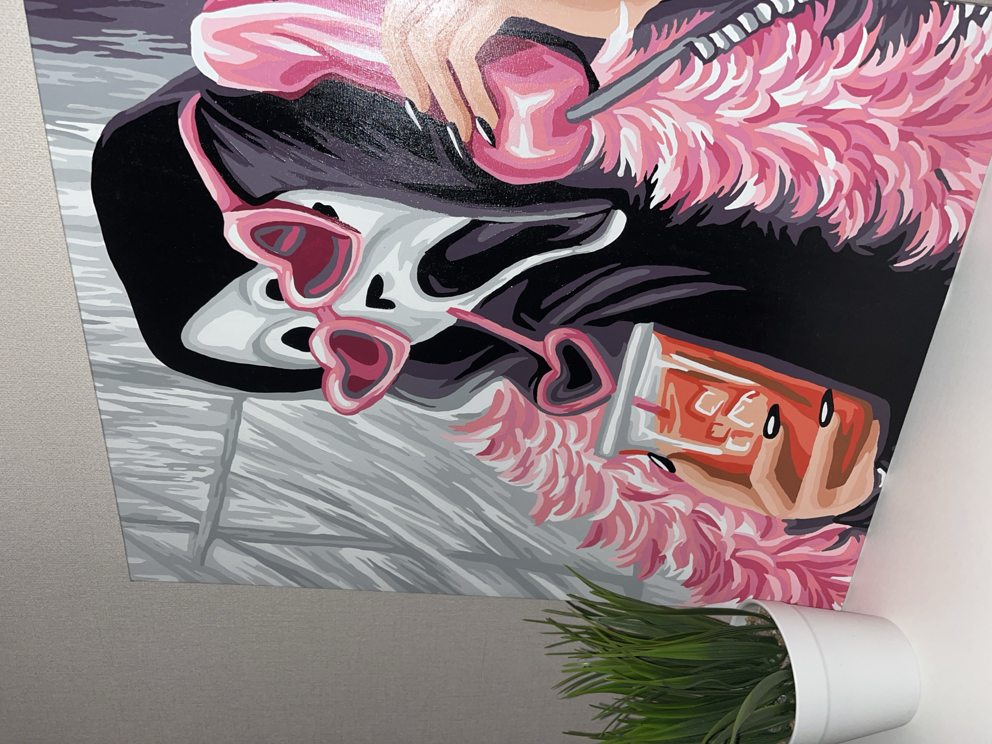 A framed painting depicting a stylized portrait is displayed, positioned near a white wall and a small potted plant. The painting showcases a human hand holding a cherry on a stick, adorned with pink feather accents. A pair of heart-shaped sunglasses rests on a surface in the painting, alongside a drink in a square glass. The background is a combination of black, grey, and white tones, creating a flat, graphic style. The plant in the lower right corner is green and appears artificial. The wall behind the artwork is light grey. This image was not taken in Krasnodar, Russia.