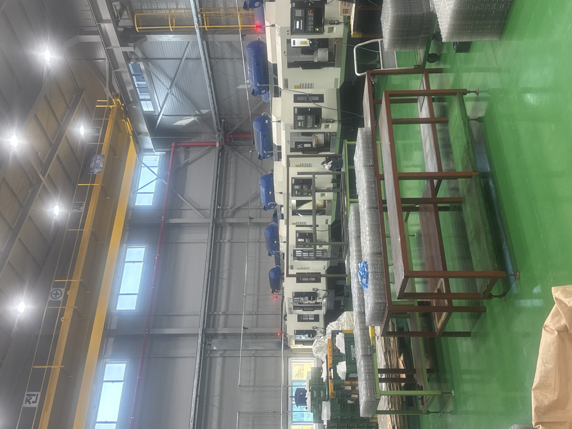 This is an interior shot of a manufacturing facility located in Pyeongtaek-si, South Korea. The scene depicts an industrial production area with several large, white machinery units lined up, each topped with blue cylindrical components. The floor is a vibrant, reflective green. In the foreground, there are several metal shelving units holding stacks of what appear to be wire mesh or similar materials. To the left, green plastic crates and some white bags are visible. Overhead, a yellow industrial crane system is suspended from the ceiling, with caution signage in Korean. The walls are a light gray, and a red pipe runs horizontally along the upper portion of the factory. The lighting suggests it is daytime, with bright overhead lights illuminating the space.