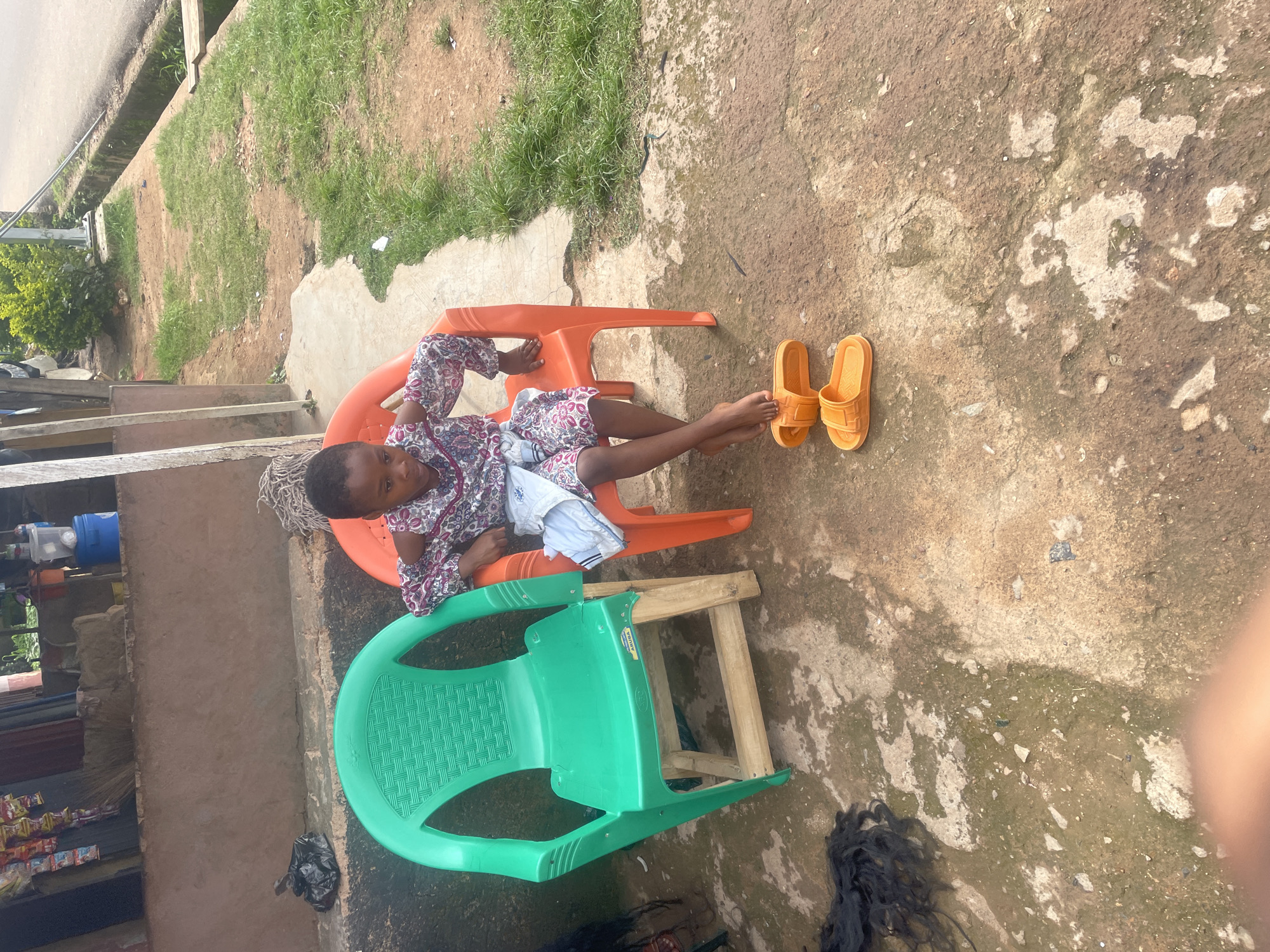 In Barakin Lerawi, Nigeria, a young child sits on an orange plastic chair. The child is wearing a patterned dress and has a cloth draped over their lap. In front of the child, on the ground, are a pair of orange slip-on sandals. To the left of the orange chair is a green plastic chair with a wooden base. The ground is a mixture of concrete and dirt, with patches of grass. In the background, there is a building with items displayed for sale and a glimpse of a road with a drainage ditch. Black hair extensions are visible on the ground to the left of the green chair.