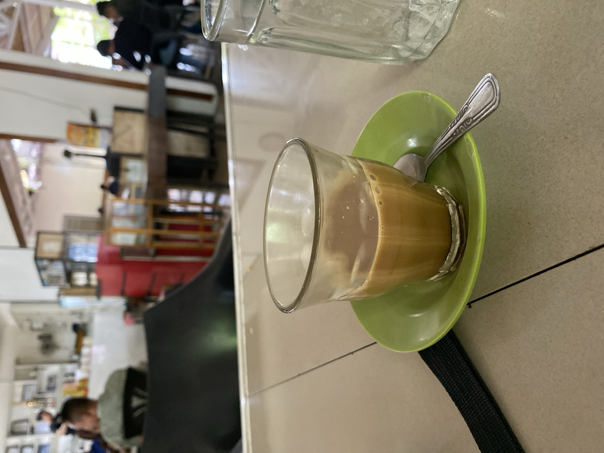 The image, taken in Lhokseumawe, Indonesia, captures an intimate view of a drink in an indoor setting, likely a cafe or eatery. The photograph is oriented vertically, rotated 90 degrees counter-clockwise.
In the foreground, occupying the lower right portion of the frame (when viewed upright), a clear glass mug holds a creamy, light brown beverage, most likely coffee or tea with milk. The mug sits on a vibrant light green saucer, beside which a silver spoon with decorative etching on its handle is placed. A black woven strap, possibly from a bag or camera, is visible beneath the saucer on the light-colored, tiled surface. Partially visible in the upper right (when upright) is another clear glass, taller and empty, displaying water spots.
The background is intentionally blurred, providing a sense of depth and activity without distracting from the main subject. It reveals an indoor space with several indistinct figures of people. Some appear to be seated, while others are moving around, suggesting a lively but casual environment. Elements of furniture, shelves, and possibly framed items or decorations are discernible, along with a striking red section of a wall or cabinet in the mid-left background. Bright natural light filters in from an unseen window or opening, illuminating some greenery outside, indicating it is daytime.
The overall scene depicts a moment in a busy yet relaxed cafe, with the focus on a beverage awaiting consumption amidst the daily life of its patrons. No discernible text is visible.
