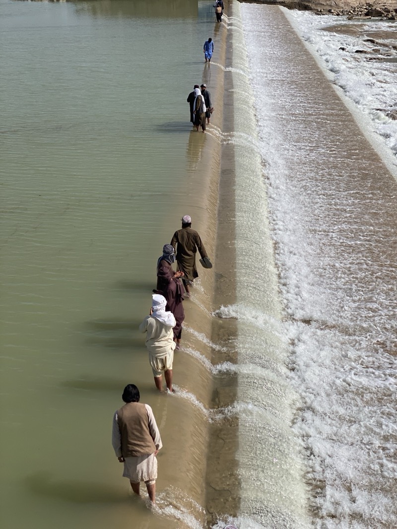 The image depicts a group of people walking across a concrete dam or weir in a river or large stream. The water appears murky and light brown in color.
People:
* There are at least nine individuals visible.
* They are dressed in traditional Afghan clothing, including long shirts, vests, and head coverings (turbans or skullcaps).
* Most are walking barefoot or with their feet in the water that is flowing over the dam.
Objects:
* A long concrete dam or weir spans the width of the waterway.
* Water is flowing over the edge of the structure.
* The far bank appears to be sandy or rocky.
Actions:
* The people are walking across the dam in a single line.
Environment:
* The scene is outdoors in daylight.
* The landscape appears arid or semi-arid.
* The location is unspecified, but the prompt suggests it may be near Kim, Afghanistan.