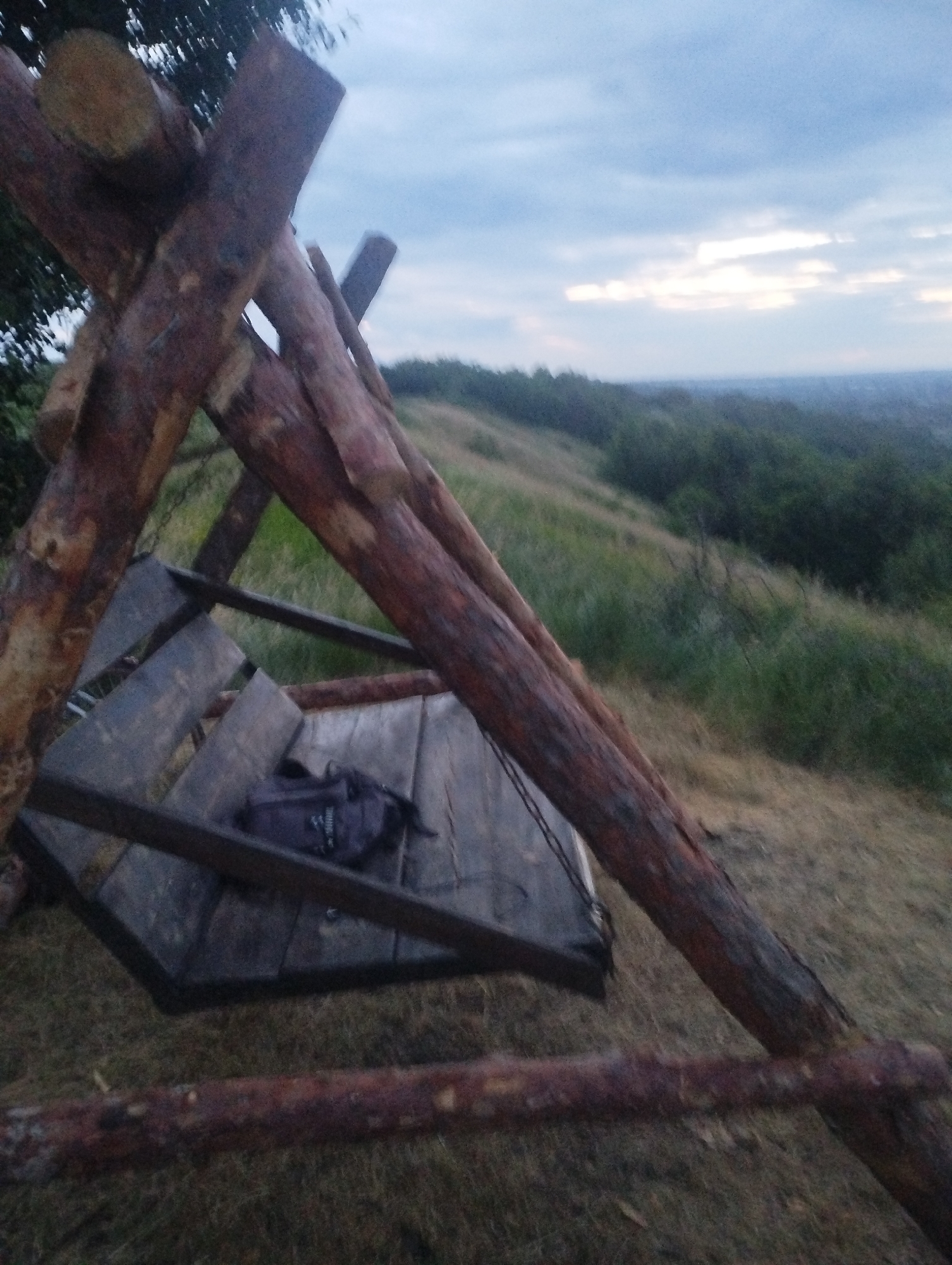 A rustic wooden swing set made of rough-hewn logs is situated outdoors. The swing set has a large, dark-colored seat made of wooden planks. A dark-colored backpack rests on the seat. The swing set is constructed with two large diagonal logs forming an 'X' shape, with additional logs supporting the structure. Behind the swing set, a grassy hillside slopes down, covered with green vegetation and trees. The sky above is overcast with scattered clouds, suggesting it may be late afternoon or evening. The location appears to be in a rural or natural setting, possibly on the outskirts of a city, with a hazy view of distant trees and buildings in the background.