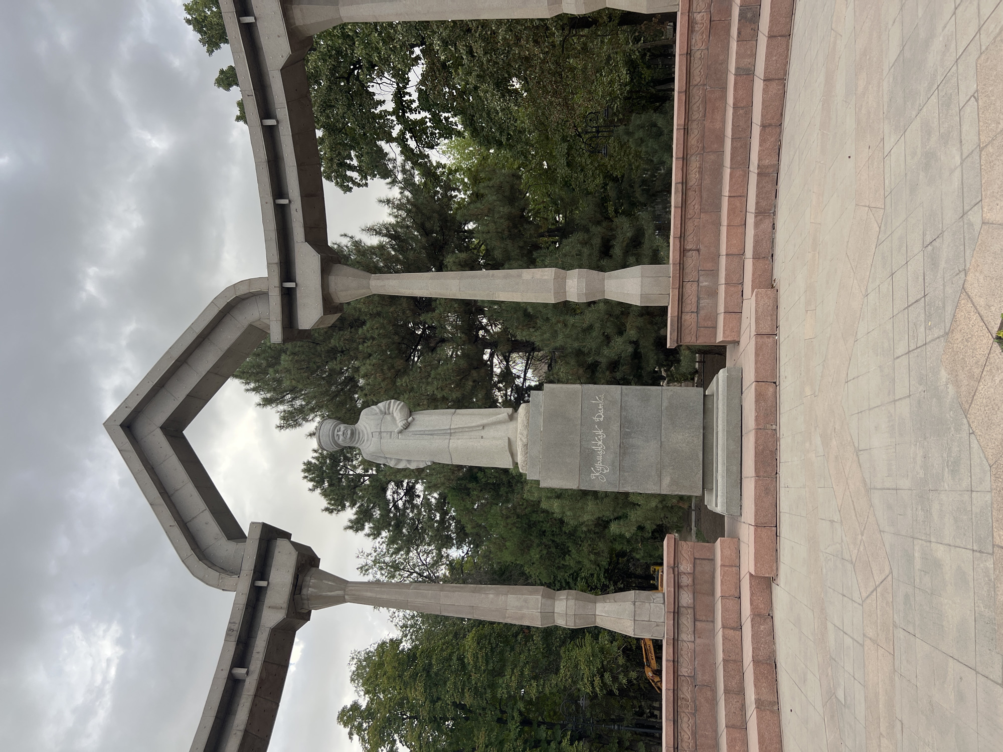 The image shows a stone monument of a person in traditional attire, standing on a pedestal in Bishkek, Kyrgyzstan. The monument is situated within an open, paved area, surrounded by trees and an overcast sky. The architecture framing the monument appears to be made of concrete pillars and beams, possibly part of a gazebo or a similar structure. The pedestal has an inscription in Cyrillic script. In the background, construction equipment, specifically a yellow excavator, is visible behind the monument, suggesting ongoing work or maintenance in the area. The ground is paved with square tiles of varying colors, arranged in a patterned manner. There are tiered, red-stone steps leading up to the monument's base. The overall scene conveys a public monument in a park-like setting with elements of urban development.