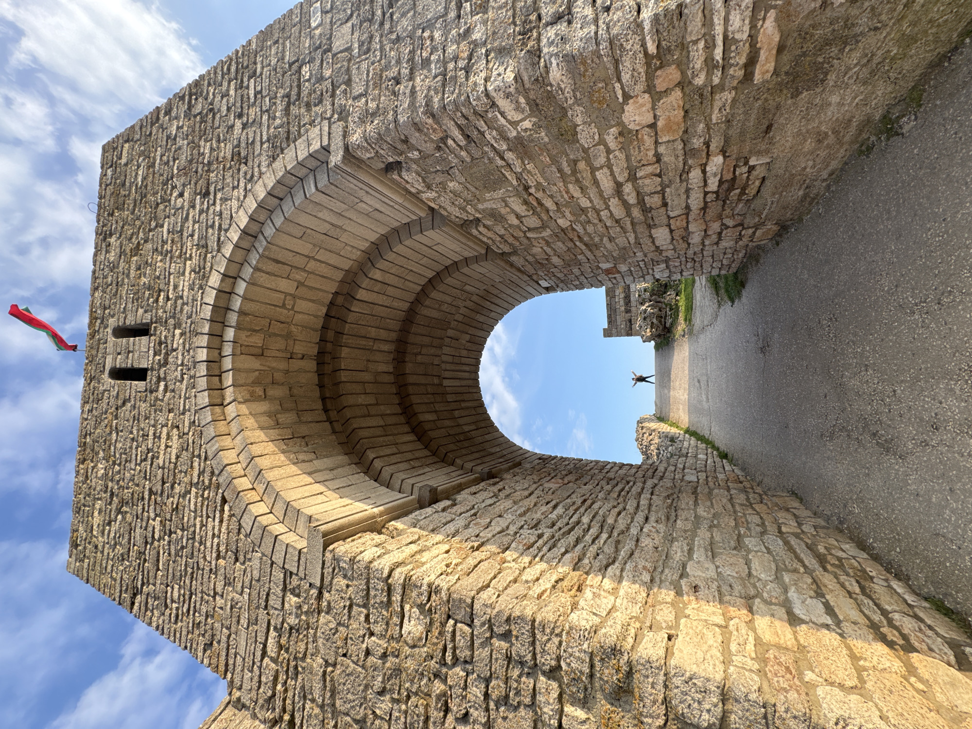 The image is a low-angle view through a stone archway. The arch is made of rough-hewn stones and constructed in a semi-circular form, leading to a pathway outside. The archway appears to be part of a larger stone structure, possibly a fortress or historic building. A person with outstretched arms stands in the distance on the pathway, under a blue sky visible through the arch. The pathway appears to be made of asphalt. A flag is visible on the left side of the archway. The location is identified as Balgarevo, Bulgaria.