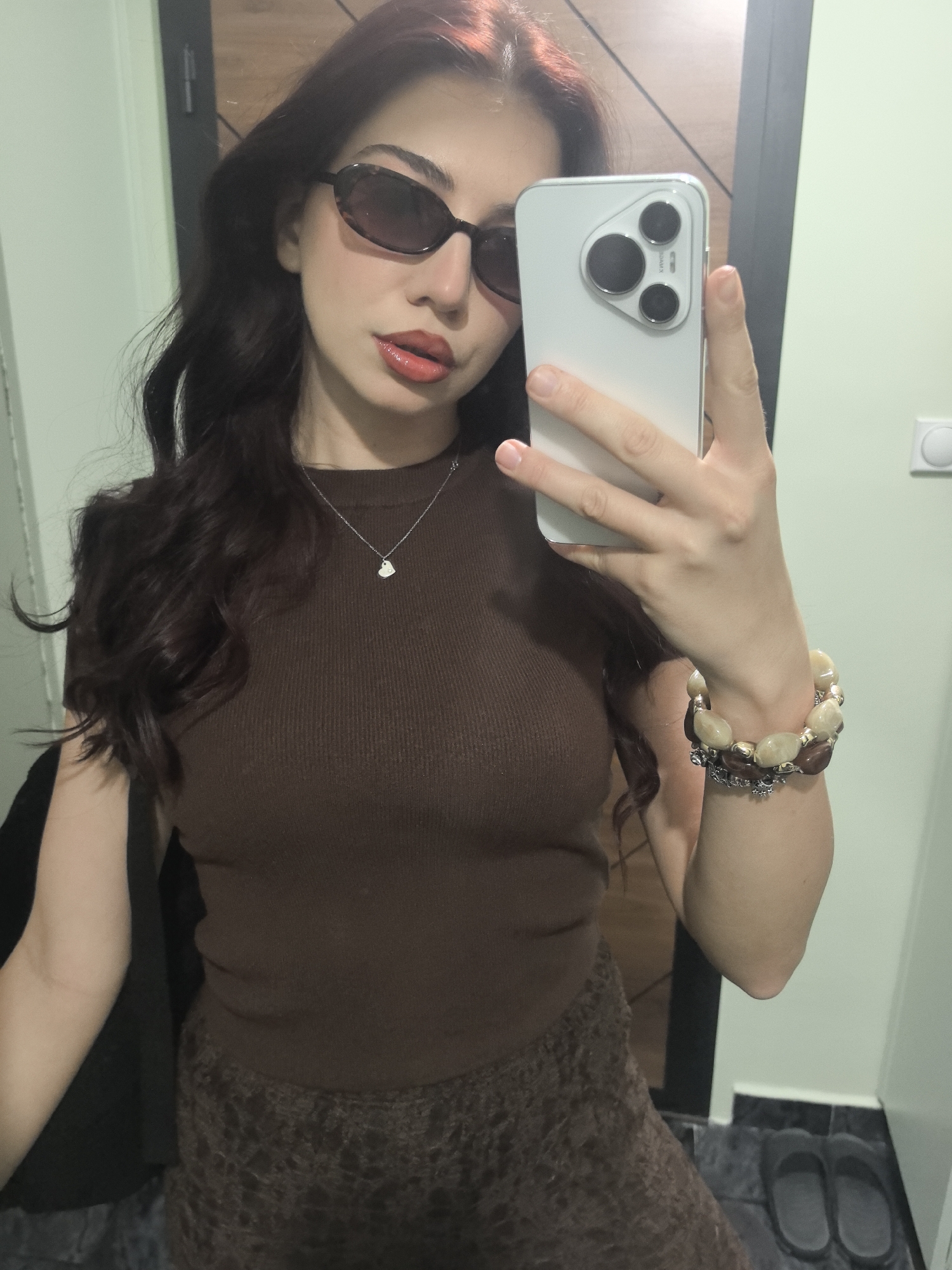 A woman wearing sunglasses and a brown top takes a selfie indoors. She is holding a smartphone in her right hand, showcasing its camera. She wears a bracelet on her right wrist and a necklace. The background includes a door and wall, along with slippers on the floor. The location is Unknown, Unknown.