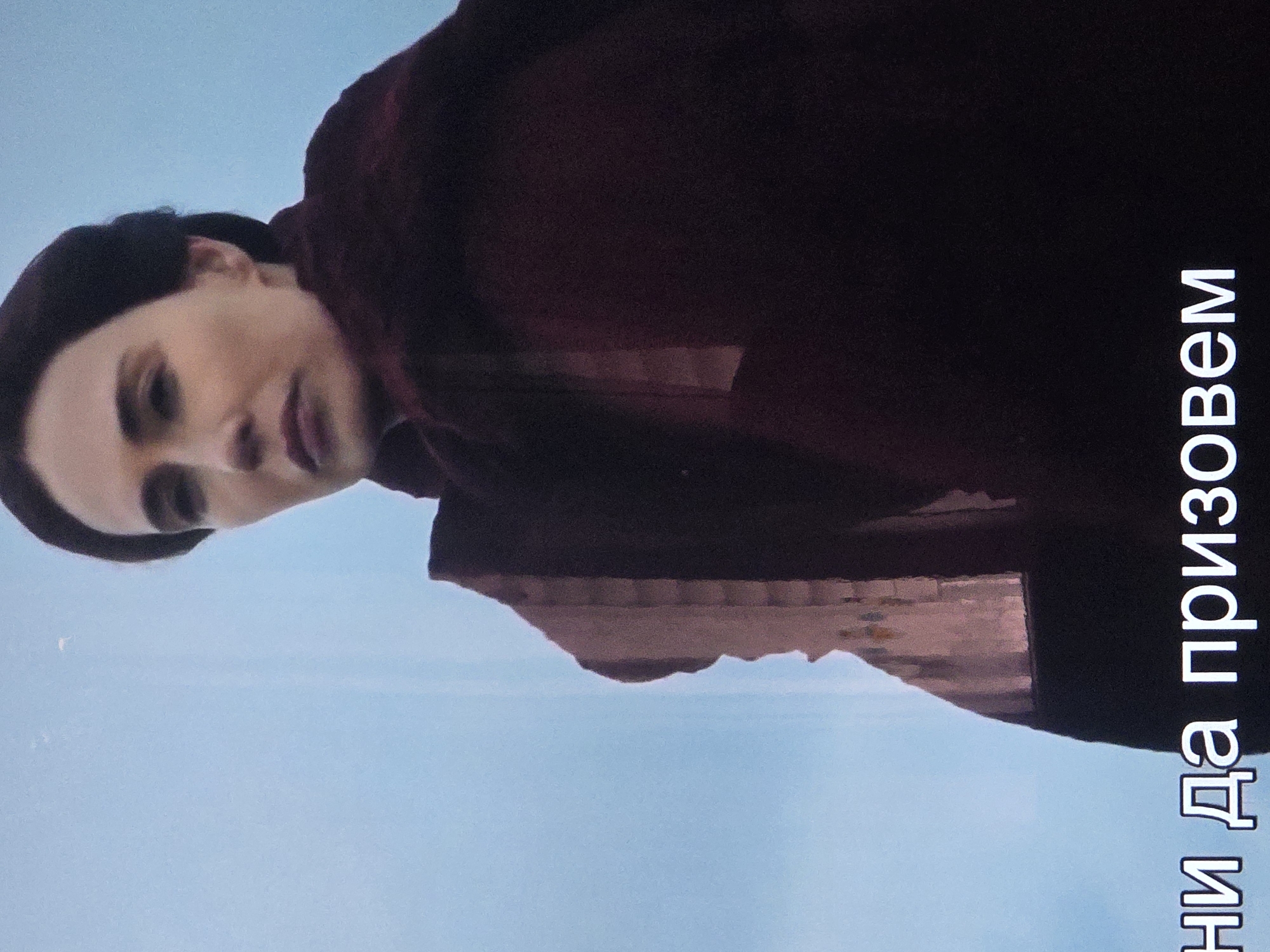 A medium shot shows a woman from the chest up, looking to the left with a neutral expression. She has dark hair pulled back and is wearing a dark red cloak. Behind her, a large, light-colored building with visible architectural details is partially obscured. In the lower part of the frame, white text in Cyrillic letters reads "ни да призовем". The background is a hazy blue sky.