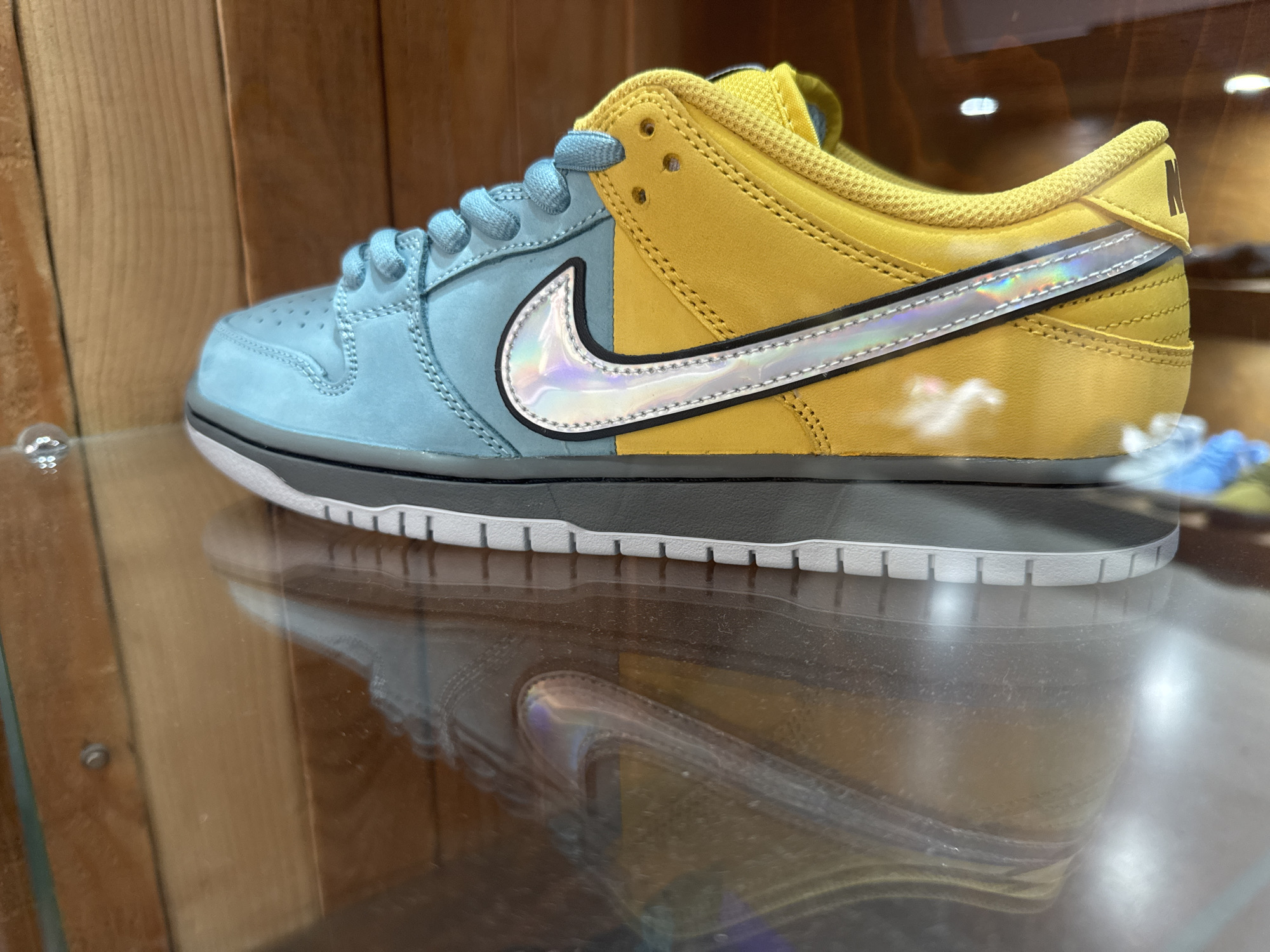 A blue and yellow Nike Dunk Low sneaker sits on a reflective glass surface inside a display case with wooden paneling. The front half of the shoe is light blue, while the back half is yellow. A silver swoosh with a black outline divides the colors. The shoelaces are blue. The midsole is dark gray and white. The reflection of the shoe is visible on the glass surface. There are no people present in the image. The location is unknown and is not possible to determine from the information provided.