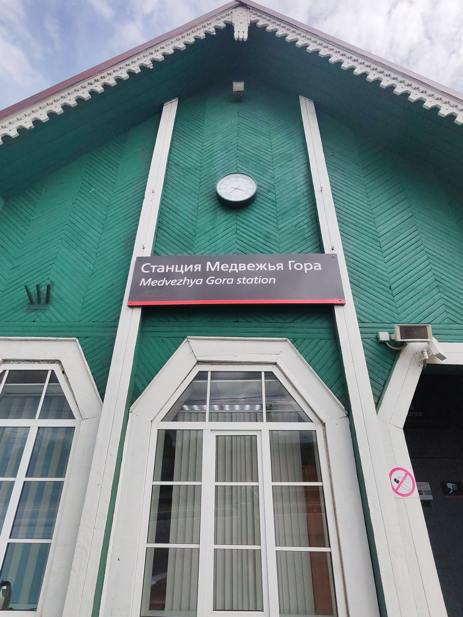 An upward-angled view captures the facade of a wooden building, identified as the Medvezhya Gora station in Medvezhyegorsk, Russia. The building is painted a distinctive deep green, with its clapboard siding laid in a diagonal pattern, creating a herringbone effect. Ornate white trim, reminiscent of gingerbread scrollwork, decorates the eaves of the gabled roof, which is visible against a bright, overcast sky.
Centrally located on the green wall is a prominent rectangular sign with a dark background and white text. It displays "Станция Медвежья Гора" in Cyrillic script above "Medvezhya Gora station" in English, with a thin red line separating the two languages. Above this sign, a round white clock with Roman numerals keeps time, though the exact time is not clearly discernible.
Below the main sign are two large, multi-paned windows with arched tops, framed in white trim that matches the architectural details. Interior blinds or curtains are visible through the windowpanes. To the right of the right window, a security camera and what appears to be a motion sensor are mounted. A circular "no smoking" sign, depicting a crossed-out cigarette within a red circle, is affixed to the white trim near a dark entrance area. A partial word, "ance" (likely part of "entrance"), can be seen on the dark surface to the far right. The overall impression is of a well-maintained, traditional station building on a cloudy daytime.