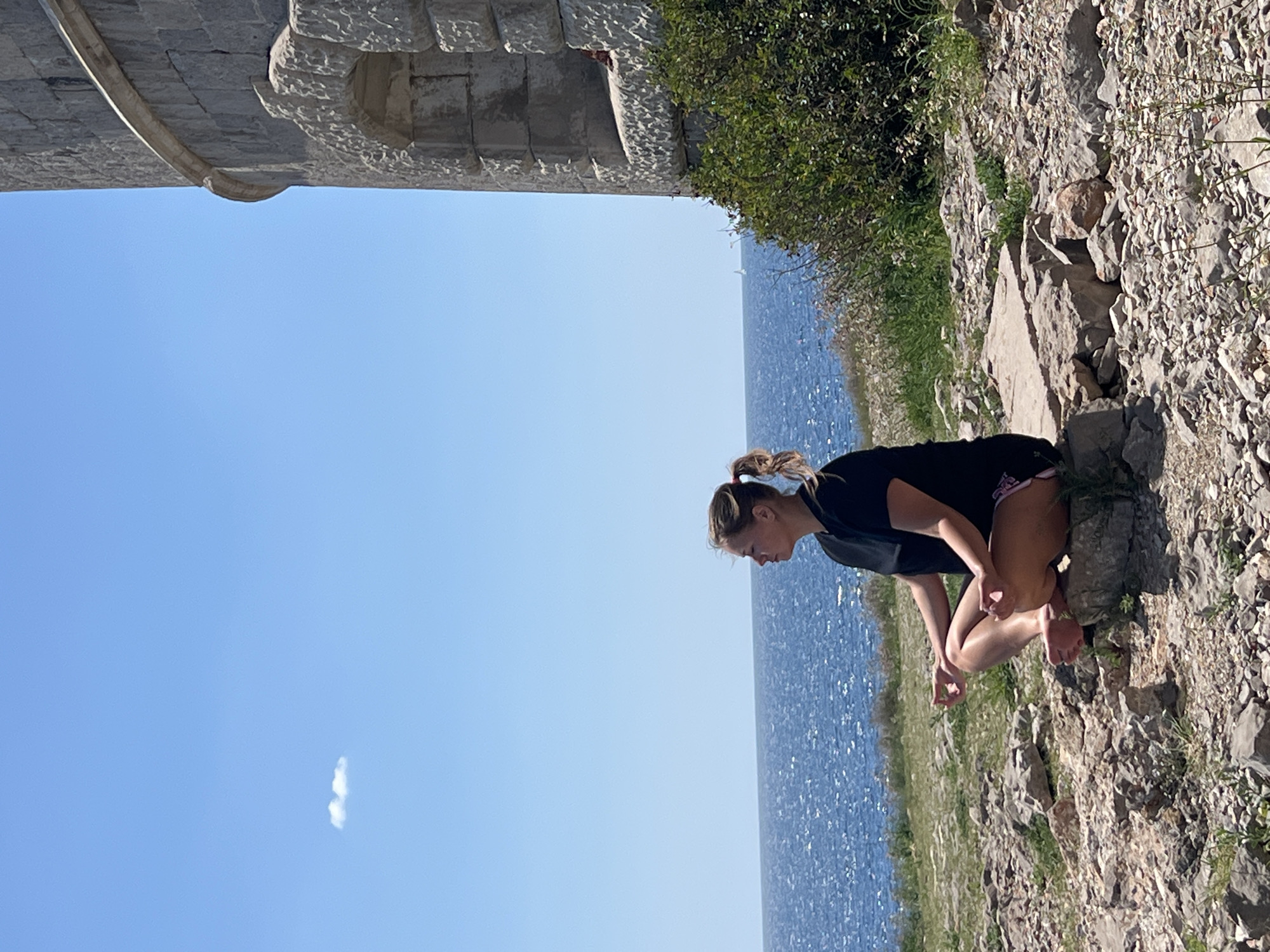 The photo shows a woman crouching near the coastline of Luštica, Montenegro. She has blonde hair in a ponytail and wears a black shirt and dark shorts with pink detailing. She is barefoot and leans forward, looking towards the sparkling blue Adriatic Sea. The rocky terrain is covered with sparse vegetation. Above her is the ancient stonework of what appears to be a fortified structure or wall. The sky is clear blue with a small, thin cloud visible.