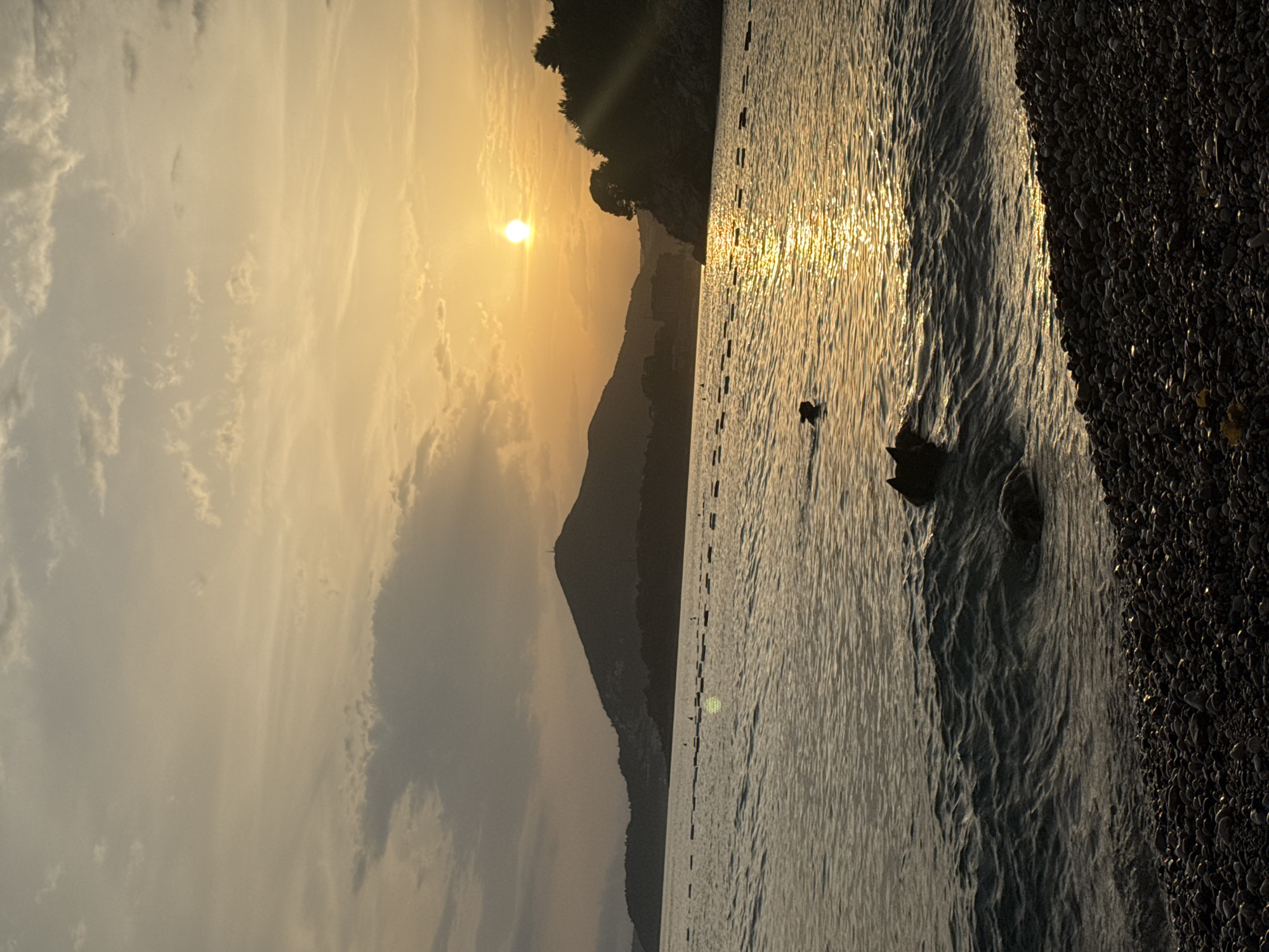 An outdoor, vertical, medium shot shows the sunset over the sea at Kamenovo, Montenegro. The sun, a bright orb, is positioned in the upper right quadrant of the frame, casting a golden sheen across the rippling water. Silhouetted against the sunset are a mountain range on the left and a forested headland on the right. The foreground is a pebble beach. Two dogs are visible in the water, appearing to swim or play. A line of buoys stretches across the water in the mid-ground. The sky is partly cloudy, with the clouds tinged orange and yellow by the setting sun.