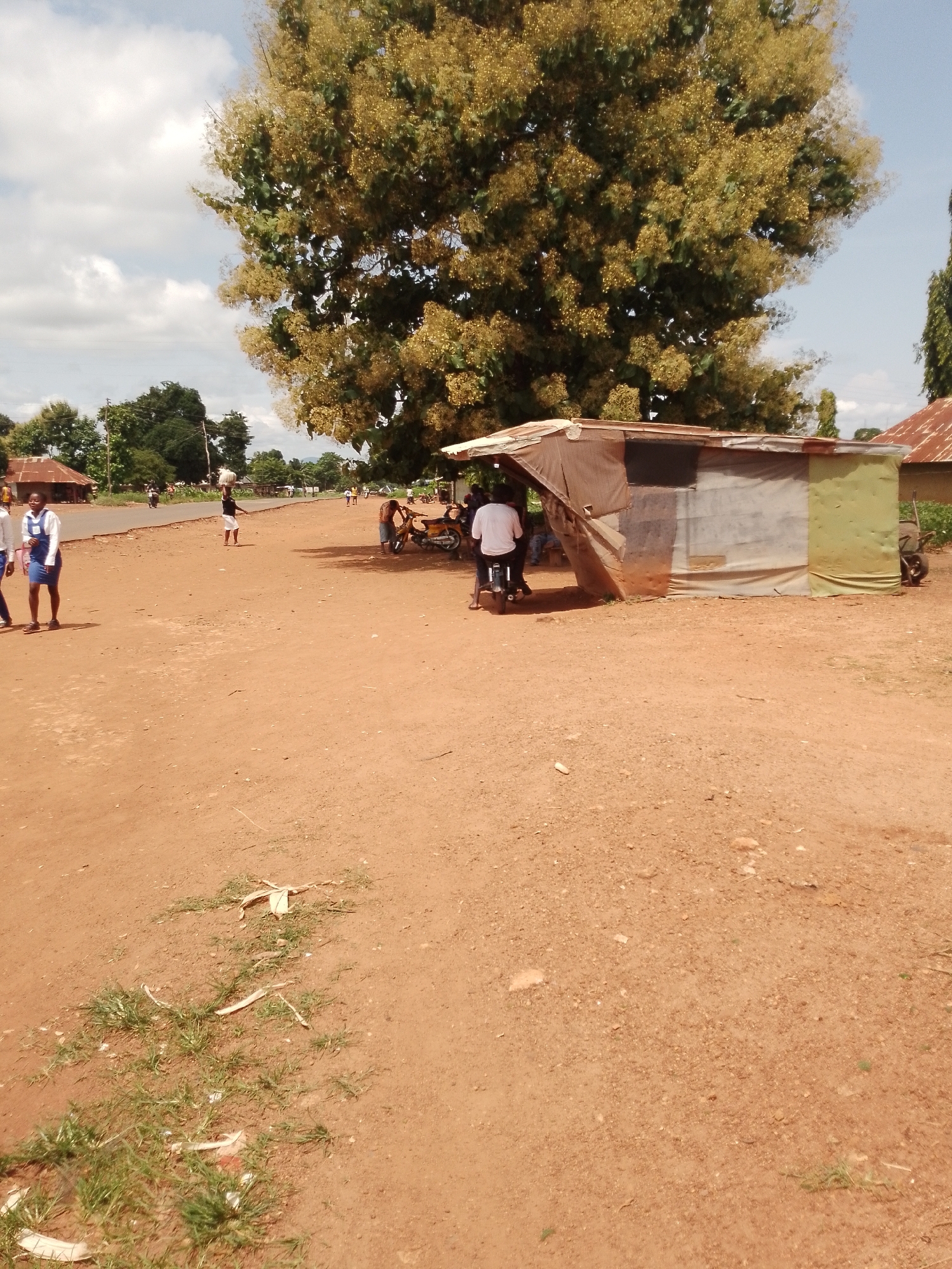 The image depicts a rural roadside scene in Adikpo, Nigeria. The ground is unpaved and reddish-brown. A small, makeshift stall constructed from corrugated metal and fabric is situated on the right side of the frame. A person wearing a white t-shirt is seated on a motorcycle parked in front of the stall. To the left, two young women in school uniforms, a white blouse and a blue dress, are walking. In the background, there are several buildings with corrugated metal roofs, trees, and people walking on a paved road. The sky is partly cloudy.