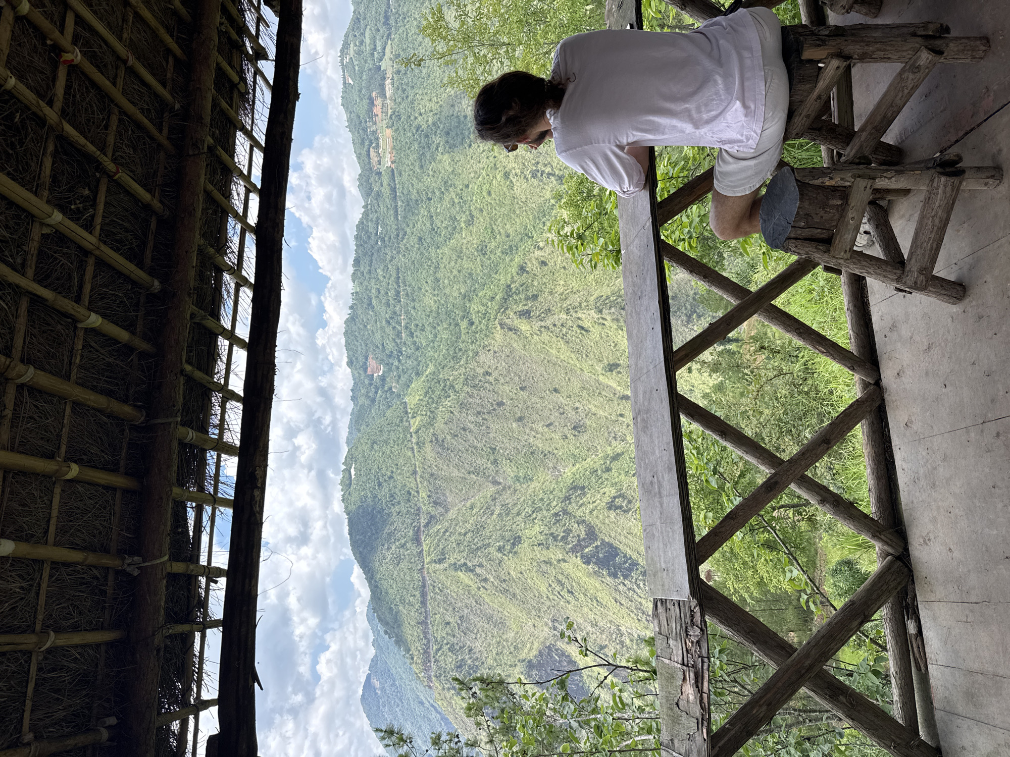 A person with dark hair, wearing a white shirt and light shorts, sits on a wooden stool, leaning against a wooden railing. The person appears to be looking out over a lush green mountain range. The foreground shows the interior of a building or structure with a thatched roof. The view extends to a distant mountain range under a partly cloudy sky. This scene takes place in Lũng Cú Commune, Vietnam.