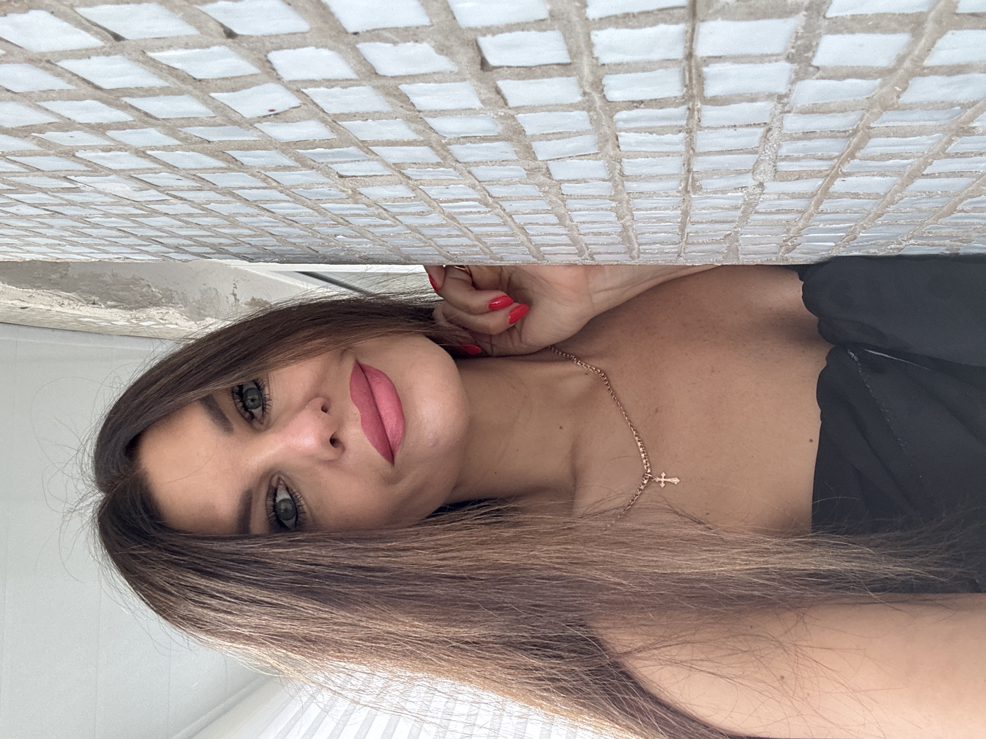 A woman with long brown hair and bright pink lipstick poses for a selfie, leaning against a tiled wall. Her left hand, with bright red nail polish, rests on the wall. She is wearing a black strapless top and a delicate gold necklace with a small cross pendant. The background is a white tiled wall with visible grout lines, and to the left, a window with blinds. The overall impression is of a casual, indoor portrait. The location is not specified in the image.