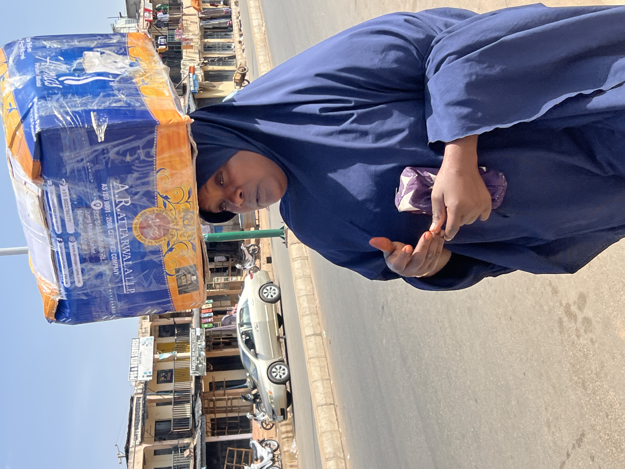 A woman in a dark blue head covering and robe balances a large, wrapped box of "A.R.ATTARWALA LLP" goods on her head. She holds a purple and white bag in one hand and gestures with the other. The setting appears to be a city street, likely in Jos, Nigeria, as referenced by the text on the box. In the background are buildings, parked motorcycles, and a light-colored car. Advertisements and signs are visible on the buildings. The scene is lit by bright daylight.