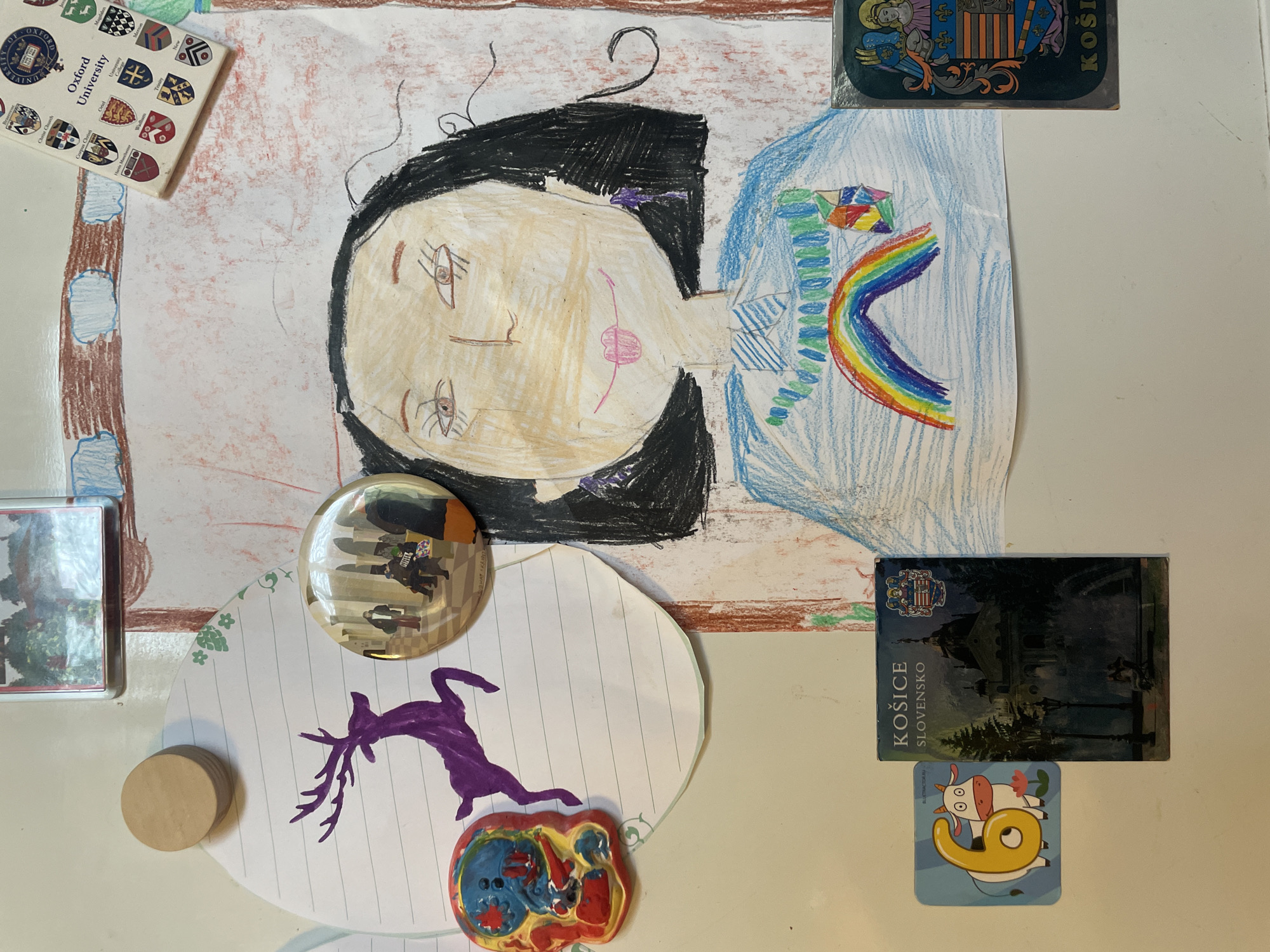 This image shows a collection of items attached to a white surface, possibly a refrigerator. These items include:
* **Drawings**: A crayon drawing of a woman with black hair, a pink cheek, and a rainbow is prominently displayed. A deer is drawn in purple marker on lined paper.
* **Souvenirs**: Magnets or souvenirs from Oxford University and Košice, Slovakia are visible. A clear round object reflects a city scene.
* **Small Objects**: A small wooden cylinder and a decorative heart-shaped object are also attached.
The items suggest a personal collection, possibly from travels and artistic endeavors. There is no indication of the city of Khimki, Russia.
