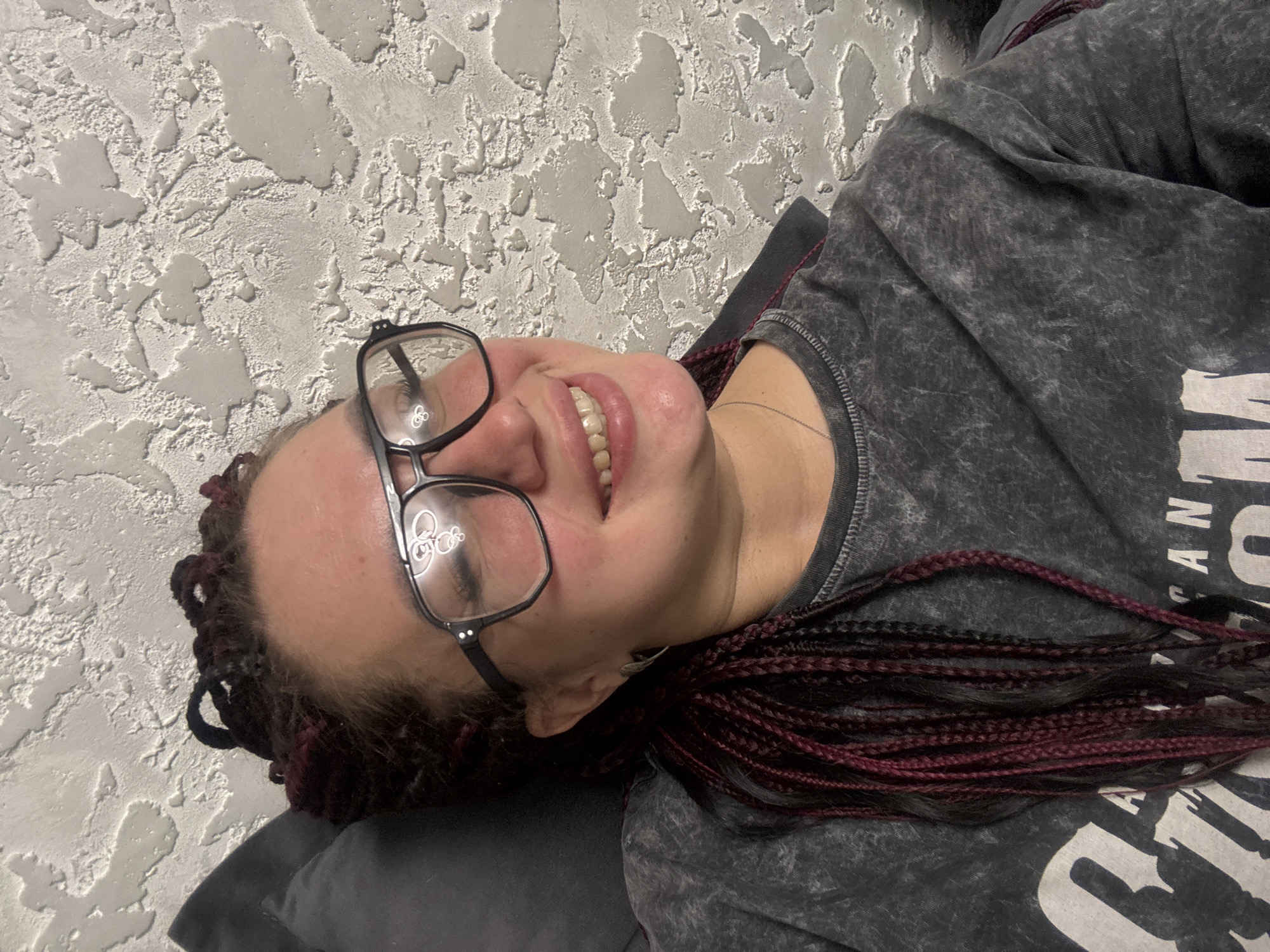 The image shows a young woman with braided hair, wearing glasses and a t-shirt. She is lying down, leaning against a pillow, with a textured wall behind her. There are no discernible elements in the image that can be used to identify the location as Tolyatti, Russia. The woman is smiling and looking away from the camera. Her hair is dark with streaks of red. She is wearing a grey t-shirt with some text on it, partially visible.