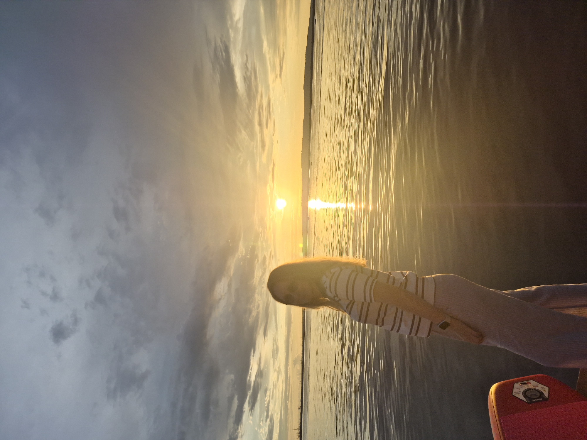 A young woman with long blonde hair and sunglasses stands on a boat, facing left and looking slightly over her shoulder towards the camera. She is wearing a striped t-shirt and light-colored pants. The setting sun casts a warm glow across a wide body of water, likely the Kama River, near the city of Chaikovsky, Russia. The sky is a mix of grey clouds and bright orange hues from the sunset. The water reflects the colors of the sky and the sun. In the foreground, a portion of the boat's red edge is visible on the left.