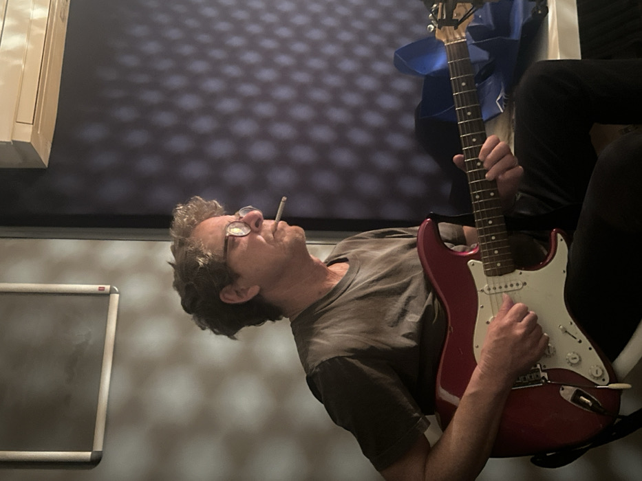 A middle-aged male individual is depicted indoors, seated, and actively playing a red electric guitar with a white pickguard. He wears round-framed glasses and has a lit cigarette between his lips. His right hand is positioned over the guitar's pickups, while his left hand frets notes on the neck. He is dressed in a dark t-shirt and dark trousers. The background consists of two distinct wall sections: a lighter section to the left with diffuse, mottled shadows, and a darker, possibly blue-purple section to the right exhibiting a repeating diamond or grid light pattern. An air conditioning unit is mounted high on the wall to the upper right. A framed object is visible on the upper left wall, and a blue object is partially visible on a surface to the man's right. The scene is located in the city of Paris, France.