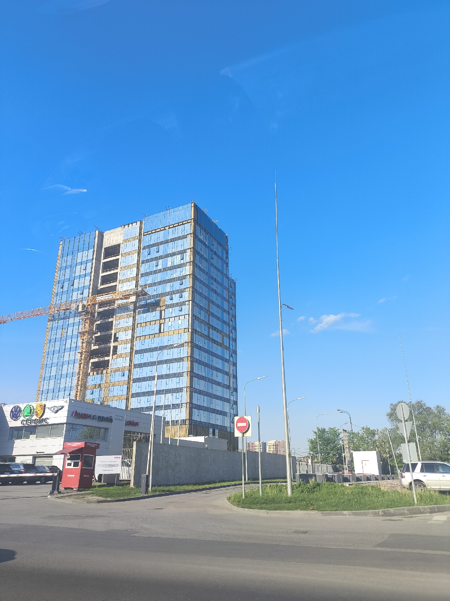 The image captures a daytime urban scene in Unknown, Unknown, under a clear blue sky. A modern high-rise building, partially covered in reflective blue glass panels, dominates the left side of the frame, with a construction crane visible alongside it. Adjacent to the building is a lower structure with automotive-related signage, including what appears to be a service center or dealership featuring logos such as Audi and Porsche. A small red booth stands near the entrance to the dealership.
The foreground is dominated by a paved road. To the right of the building, there's a grassy traffic island with road signs and a white SUV parked nearby. Further in the background are apartment buildings and green trees. The overall environment suggests a commercial or industrial area on the outskirts of a city.