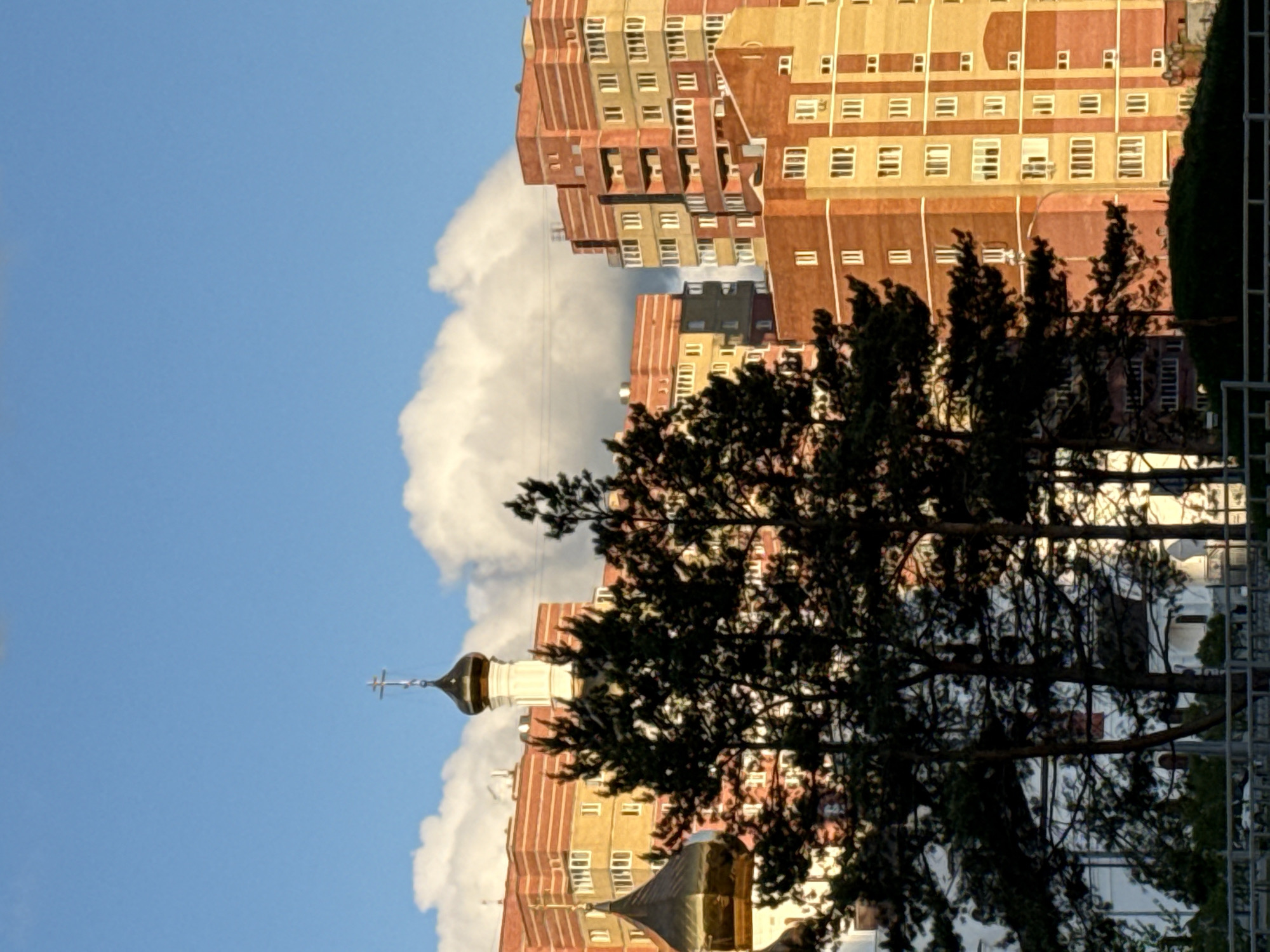 The image depicts a scene in Tobolsk, Russia, featuring a skyline juxtaposing older and more modern architecture. In the foreground, a silhouette of tree branches partially obscures the view, framing a Russian Orthodox church with a prominent golden dome and a cross atop its spire. White fluffy clouds and a blue sky form the backdrop to the left of the church, contrasting with the angular, multi-story apartment buildings of yellow and red brick to the right. These buildings exhibit a grid-like pattern of windows, suggesting a residential area. A metallic fence is faintly visible. The lighting suggests a late afternoon or early morning scene, given the angle and softness of the light on the buildings.