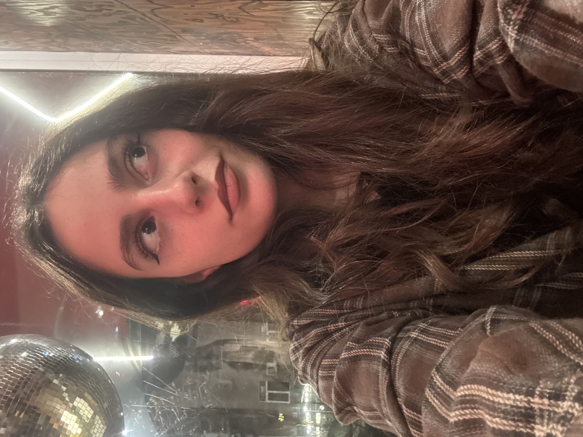 A female subject with long, dark brown hair and dark winged eyeliner is depicted in a medium close-up, gazing towards the upper right. She wears a brown and tan plaid long-sleeved shirt. Ambient lighting casts a reddish tone on her face. In the upper left background, a suspended disco ball reflects light. Behind the subject, a window or glass panel reflects an exterior urban environment, featuring a building facade with visible windows and bare tree branches. A vertical linear white light fixture and a patterned interior wall are also discernible. The scene is located in Yerevan, Armenia.