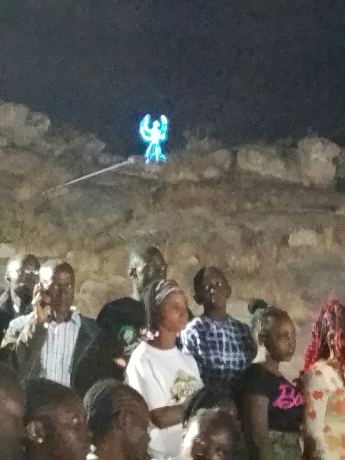 A blurry, dimly lit outdoor scene presents a crowd of dark-skinned individuals, most likely children and adults, gazing upwards. A luminous figure resembling a person with outstretched arms stands atop a rocky or cloudy formation. The overall atmosphere suggests an event or spectacle, with the bright figure serving as a focal point against the dark sky and background. Some members of the crowd are more visible, showcasing different attire like a white t-shirt, plaid shirt, and a jacket.
