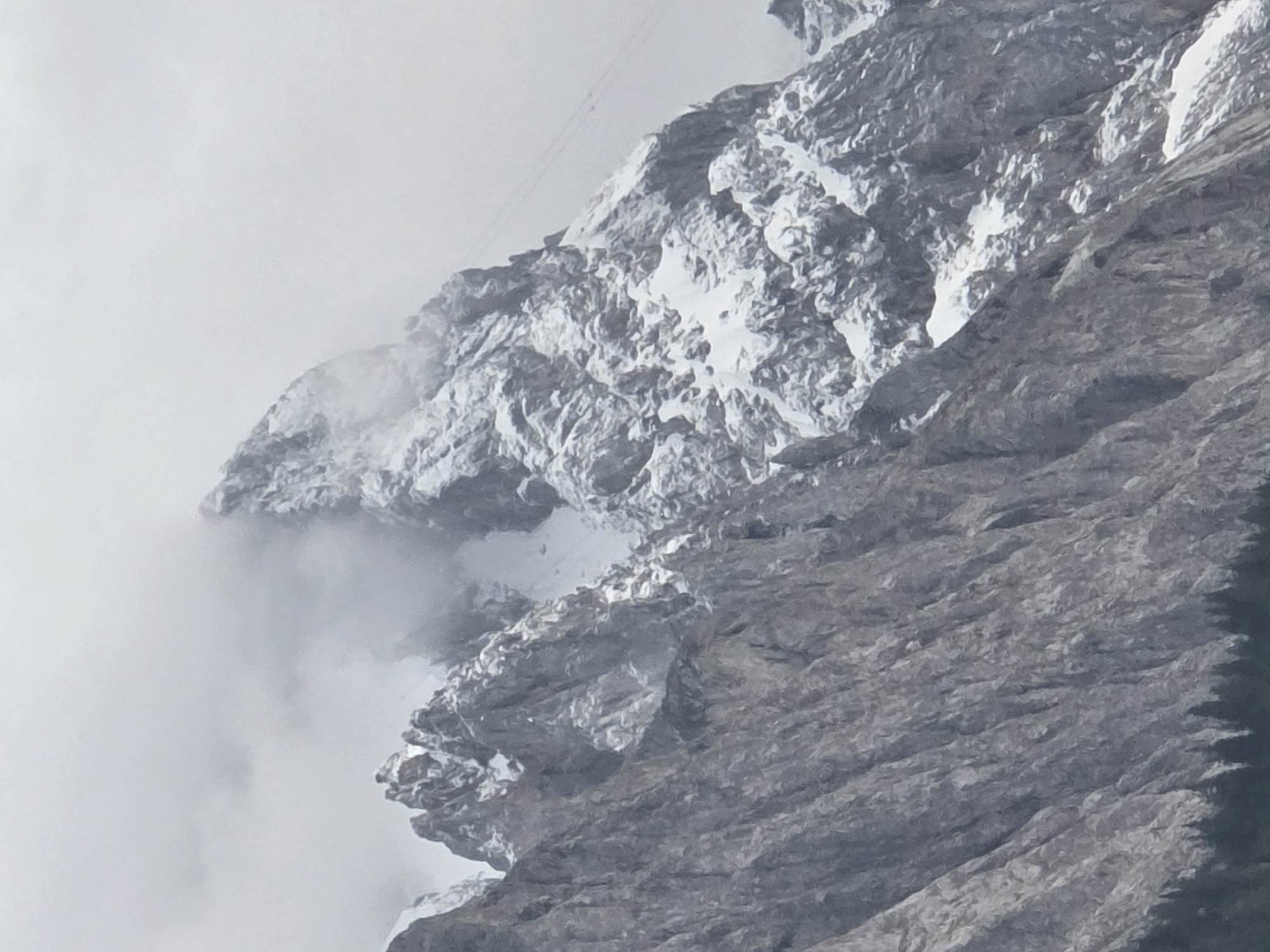The image shows a jagged, snow-capped mountain peak shrouded in mist, characteristic of the Zugspitze massif in Garmisch-Partenkirchen, Germany. The foreground is dominated by dark, craggy rock formations with sharp vertical lines, possibly indicating a steep ascent or cliff face. Patches of snow are visible on these rocks, suggesting a cold environment. In the background, the main peak is heavily dusted with snow, with swirling clouds obscuring parts of its summit and ridges. A faint, thin cable line is visible ascending the snowy slope, likely part of a cable car system for reaching the summit. The sky is overcast and a uniform grey, contributing to the dramatic and slightly foreboding atmosphere. At the very bottom of the image, a dense line of dark green coniferous trees forms the base of the mountain, contrasting with the lighter tones of the rock and snow. There are no people or man-made structures other than the cable line.