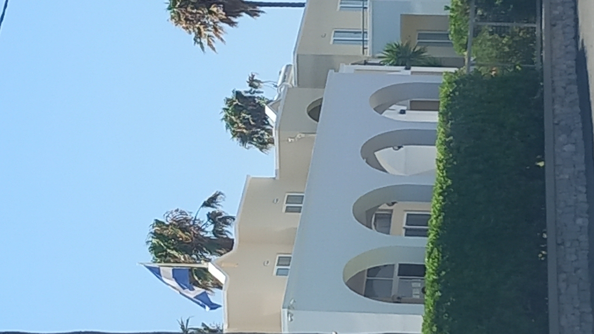 The image shows a building with a Greek flag flying from a flagpole. The building appears to be in a sunny, outdoor environment. The architecture of the building is characterized by arches and a light-colored facade. Palm trees are visible around the building, suggesting a warm climate. The image is taken from a slightly low angle, looking up at the building and the flag. The sky is clear and blue. There is a green hedge in the foreground. The scene suggests a location in Greece, possibly near the sea given the palm trees.
