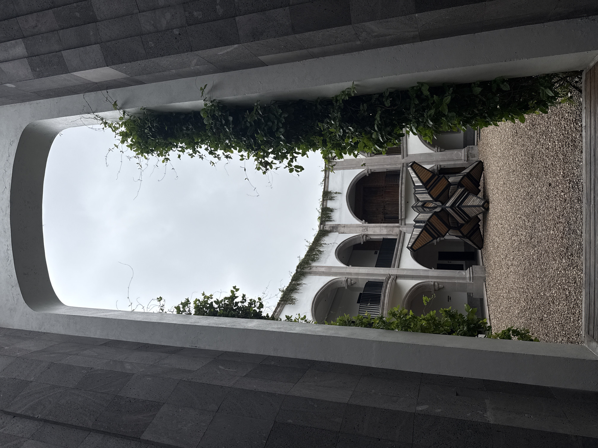 The image shows an interior courtyard with a central sculpture in San Miguel de Allende, Mexico. The courtyard is surrounded by a white building with arched balconies and is partially covered in gravel. Vines climb up the white walls and a dark stone facade frames the view. The sculpture is abstract, made of wood and metal, and resembles a butterfly or a starburst. The sky is overcast.