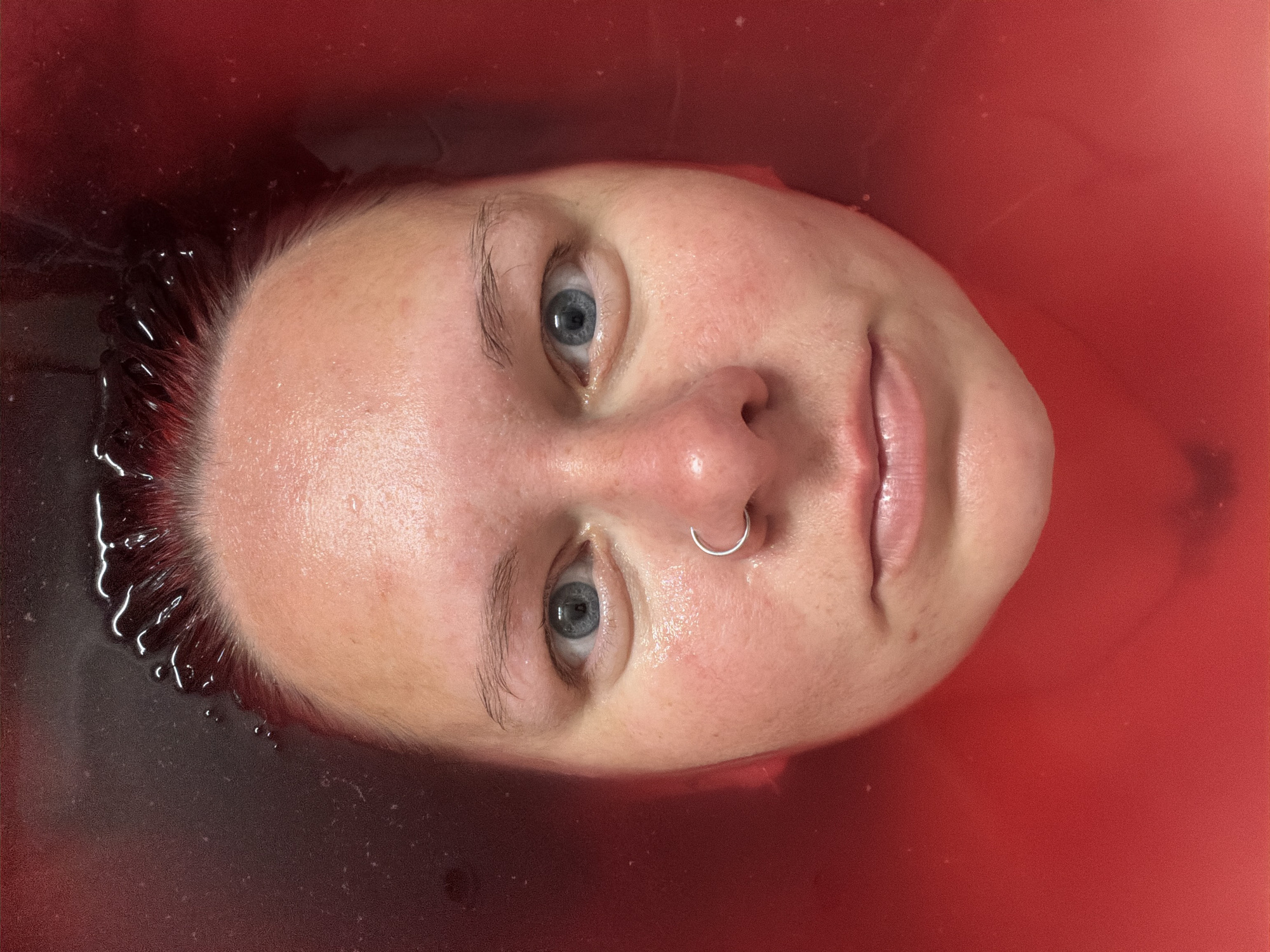 A close-up, overhead shot depicts a person's face submerged in a red liquid. The person has fair skin, light eyebrows, and blue eyes. They have a nose ring in their left nostril. The hair near the forehead appears damp and slightly darker in color, suggesting it is wet. The surrounding liquid is a consistent, dark red hue. The picture was not taken in the city of Chaikovsky, Russia.