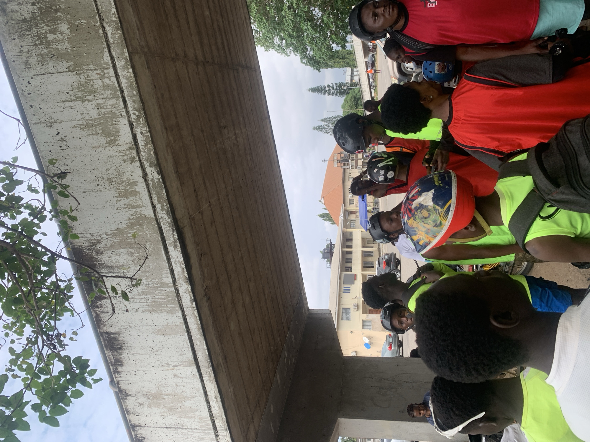A group of individuals, primarily young males, are gathered under a concrete overpass in Alagbede, Nigeria. Many are wearing colorful vests, primarily red and neon yellow, and several have helmets on their heads. Some helmets are plain black, while others are decorated with patterns and designs. The individuals appear to be looking in various directions, suggesting they might be waiting or discussing something. Behind them, a building with a cream-colored facade and orange-tiled roof is visible, along with cars and trees, indicating an urban or semi-urban environment. The overall lighting suggests it is daytime, with an overcast sky visible through the leaves of a tree extending from above the overpass. The concrete structure of the overpass shows signs of wear and tear, with water stains and some exposed rebar.