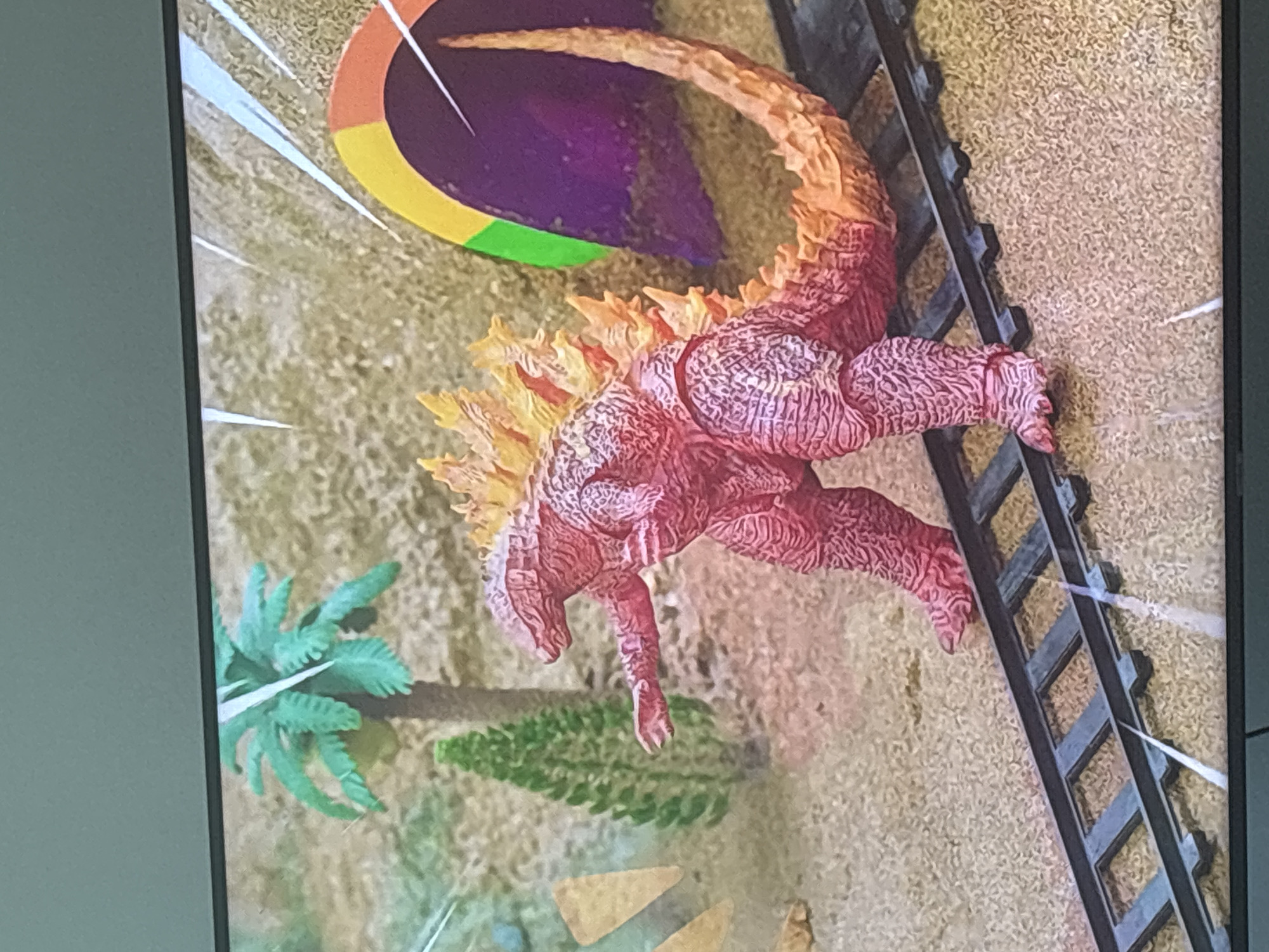 The image shows a close-up, angled view of a red Godzilla toy figure standing on a miniature train track. The background appears to be a diorama with sandy ground, a small green artificial tree, and a larger artificial palm tree. To the right, there is a portion of a colorful tunnel or archway, with segments of purple, green, yellow, and orange visible. White streaks, possibly representing motion or light effects, are present throughout the scene. The Godzilla figure has orange and yellow spikes along its back and tail. The overall impression is that of a toy display. There is no indication of any location in Dubai, United Arab Emirates.