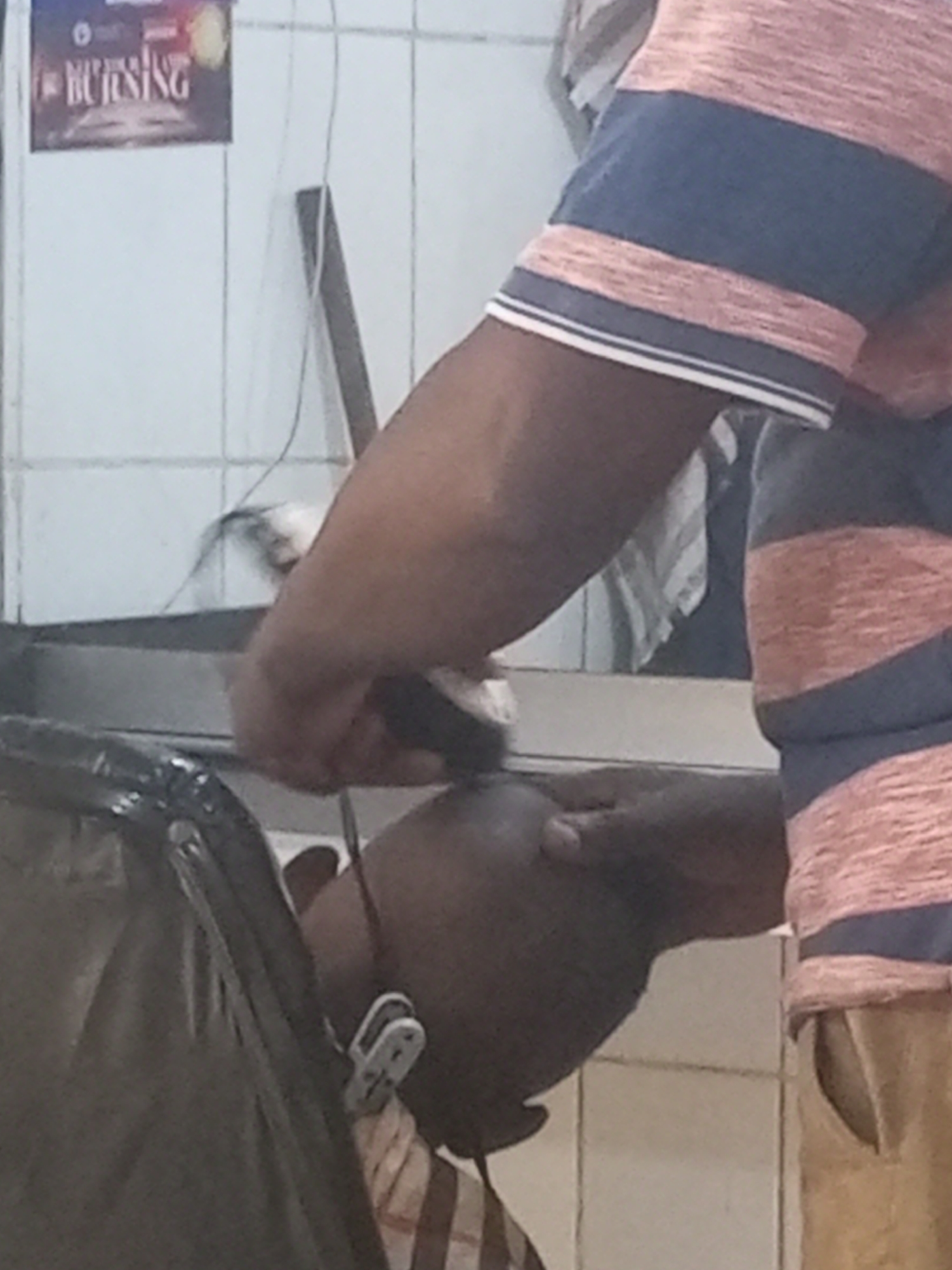 A barber is giving a haircut to a customer. The barber is wearing a striped shirt and khaki pants. The customer is sitting in a black barber chair. The location is likely a barber shop in Yelwa, Nigeria, given the poor lighting and tile walls.