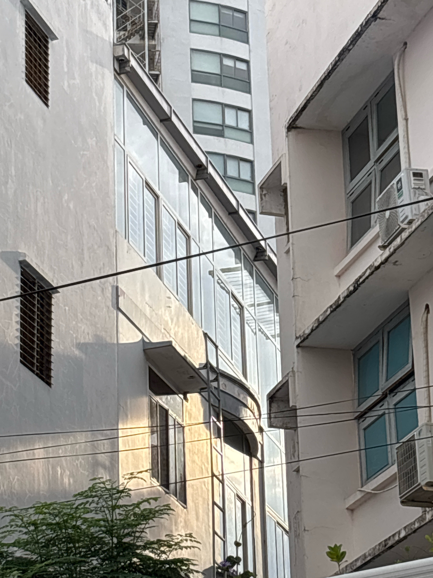This image depicts a tight urban street scene in Nha Trang, Vietnam, focusing on the architectural details of adjacent buildings. On the left, a white, textured building features a dark-grated window and a long, reflective glass facade with multiple panes. A tree with lush green foliage is partially visible at the bottom. To the right, a lighter-colored building exhibits a series of windows with light blue panes, an outdoor air conditioning unit, and a slightly weathered concrete overhang. Power lines crisscross the frame, suggesting a dense urban environment. The lighting indicates it is likely late afternoon or early morning, with warm sunlight casting shadows and highlighting textures on the buildings. The perspective is from a low angle, looking upwards.