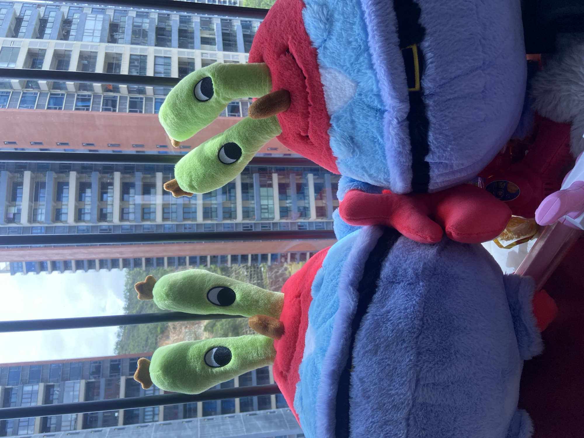 Two Mr. Krabs plush toys are positioned in front of a window with a view of modern high-rise buildings in Huizhou, China. The plush toys are colorful, with the top halves featuring green tentacles with eyes and small brown accents, and the bottom halves colored in red, light blue, and gray with a black belt detail. One plush toy is slightly to the left of the other. The window has a black metal grate in front of it. The background shows the architecture of a dense urban environment, with a mix of residential and commercial buildings, some with balconies and glass facades. The lighting suggests it is daytime, possibly overcast.