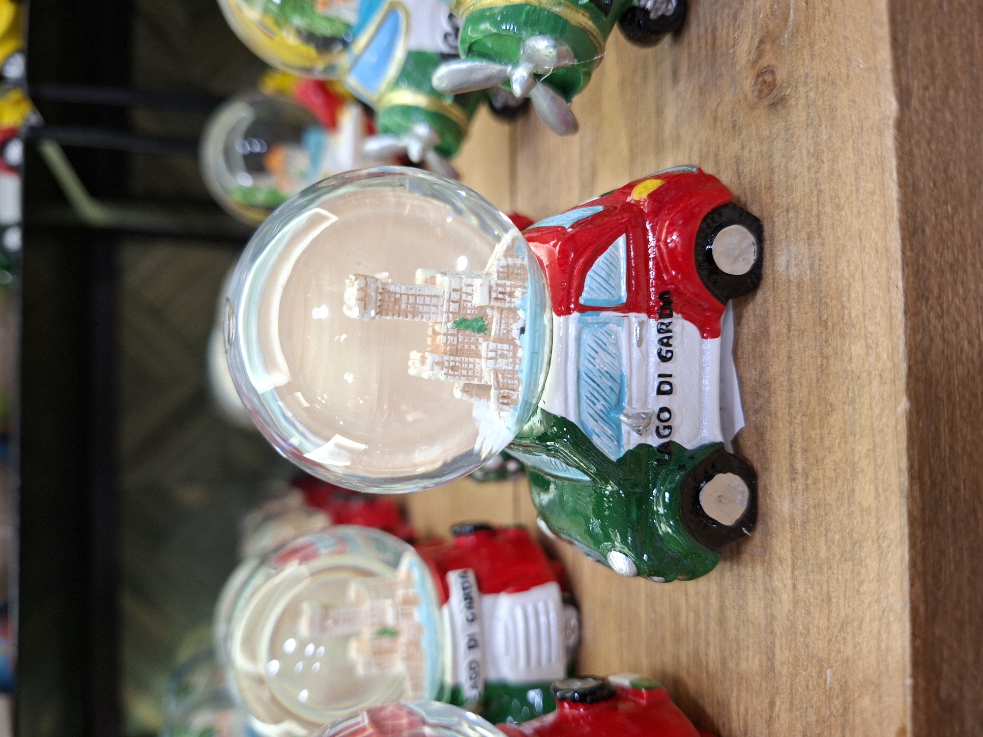 The image displays a collection of snow globes, each designed to resemble a miniature vehicle with a castle inside. The snow globe in the foreground is shaped like a small car, painted in the colors of the Italian flag: green, white, and red. The text "LAGO DI GARDA" is visible on the white portion of the car, indicating a connection to Lake Garda. Other snow globes, also featuring miniature castles and vehicle designs, are visible in the background, some appearing to be airplanes. The snow globes are arranged on a wooden surface, likely a shelf or table, with a blurred background suggesting a retail or display environment. The scene is well-lit, and the details of the snow globes are in sharp focus. The snow globes are decorative souvenir items, and their presence suggests a commercial setting, possibly a gift shop in or near Peschiera del Garda, Italy.