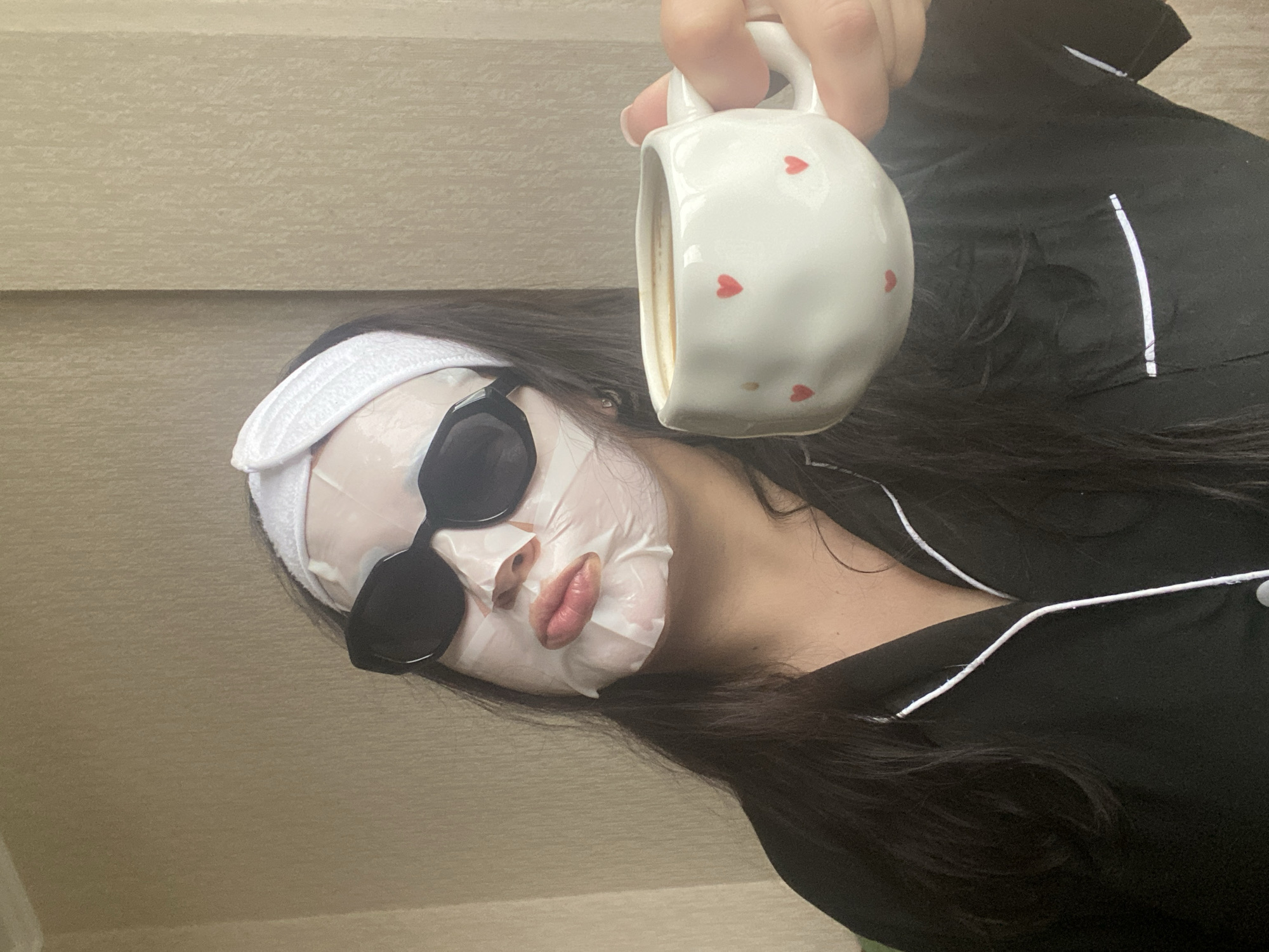 A person, facing diagonally, is wearing a white facial sheet mask and black sunglasses. They also have a white headband and are wearing black pajamas with white piping. They are holding a white mug with red hearts on it. The background is a textured wall. The image appears to be taken indoors. There is no indication of the location being Sochi, Russia.