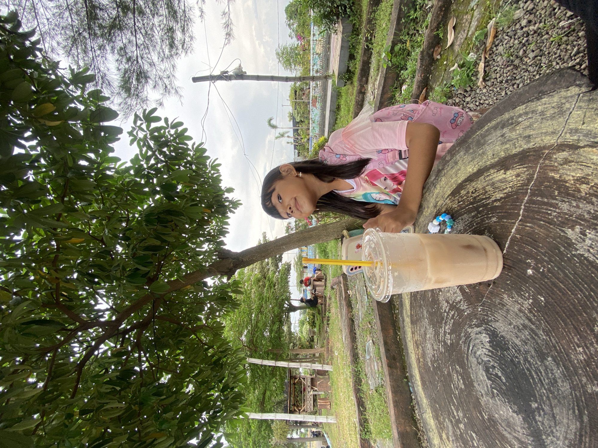 A girl in a pink shirt and black hair leans over a large, round wooden table. A clear plastic cup with a yellow straw sits on the table next to her. The table is situated outdoors, possibly in a park, in Kalitengah, Indonesia. Green trees and vegetation surround the area. In the background, there are benches and people sitting. A utility pole with wires is visible in the distance.