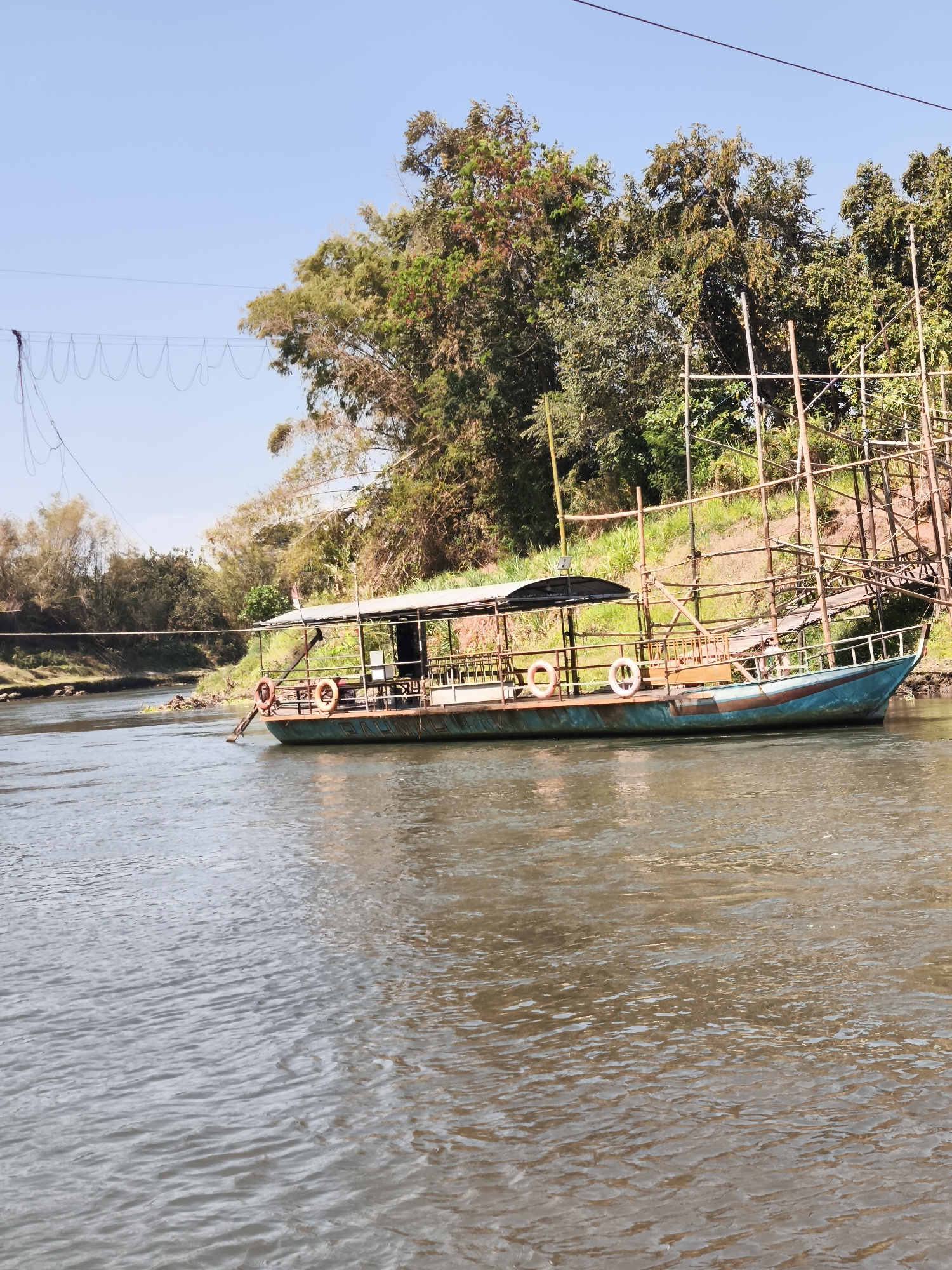 A long, narrow boat with a covered seating area is moored on a calm, muddy river. The boat is painted in teal and brown, and it has a metal railing along its sides. Two life rings are visible on the deck. In the background, a lush green, tree-lined bank rises steeply. The trees are a mix of leafy deciduous and bamboo varieties. Overhead, a thin wire is strung across the frame, attached to a series of loops, possibly part of a zipline or an old electrical wire. The sky is a clear, pale blue. The image appears to have been taken from another boat on the river. The scene is set in Papar, Indonesia.
