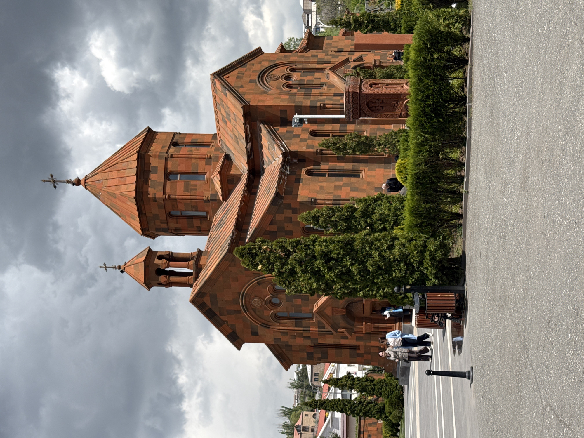A full shot depicts Saint Anna Church in Yerevan, Armenia on a cloudy day. The Armenian Apostolic church is constructed of dark orange brick in a traditional architectural style, featuring gabled roofs and a steeple topped with a cross. The church is surrounded by neatly trimmed bushes and tall trees. A paved walkway runs along the right side of the image, adjacent to the church grounds. A few people are sitting on benches along the walkway. In the background, buildings and vegetation can be seen, indicating an urban setting. The sky is overcast with dark, heavy clouds.