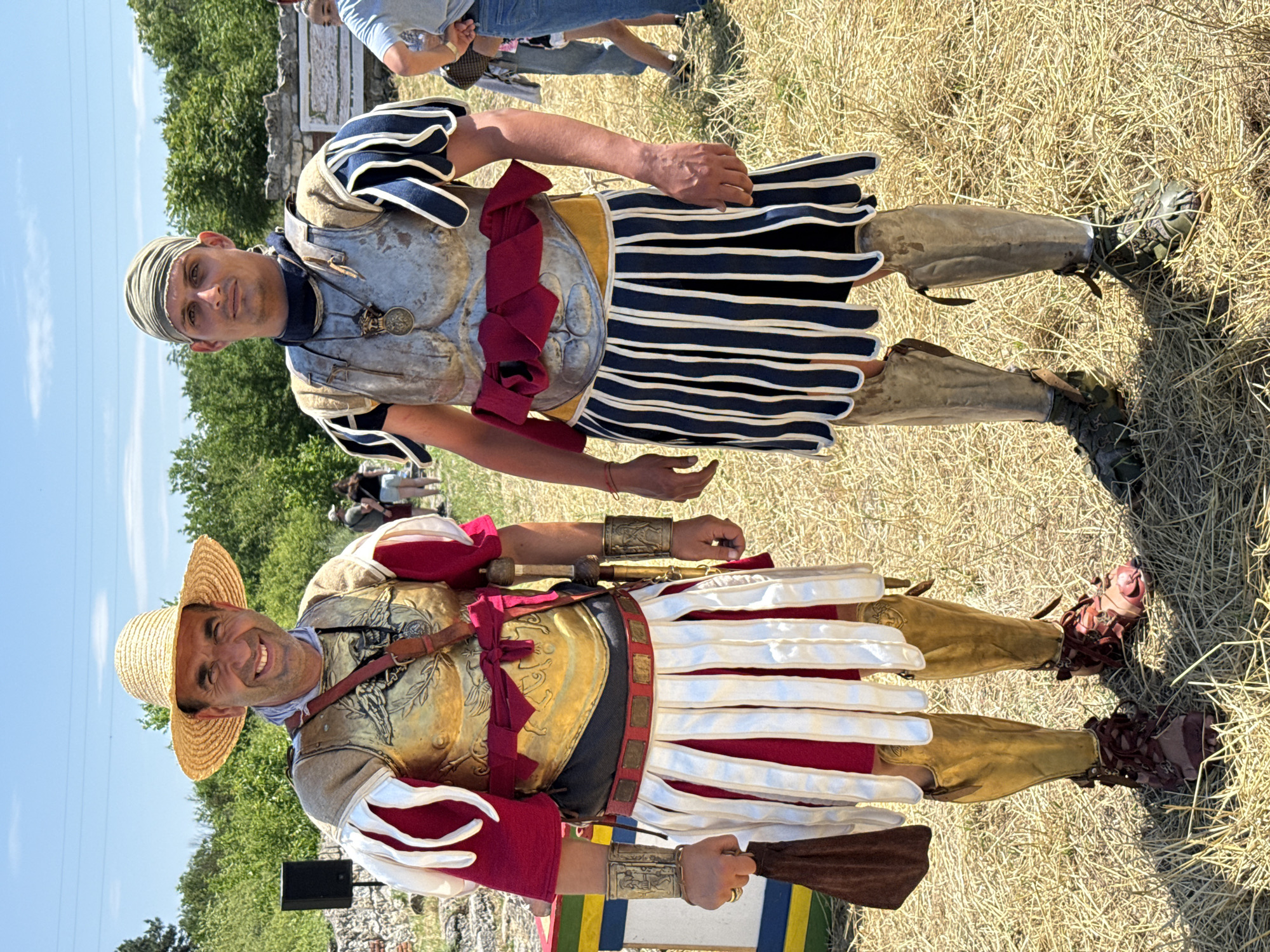 The image depicts two adult males in historical, possibly Roman, reenactment costumes, standing outdoors on a sunny day in Gigen, Bulgaria.
The male on the left, exhibiting a smiling facial expression, wears a wide-brimmed straw hat. His torso is covered by a golden cuirass, embellished with a central winged motif resembling an eagle, flanked by laurel wreaths. Beneath the cuirass, a red, short-sleeved tunic is visible, with white and red puffed shoulder elements. A dark brown leather strap with a circular medallion diagonally crosses his chest. A red sash is tied around his waist over a dark undergarment, which transitions into a white pteryges skirt with vertical pleats, revealing red fabric underneath. He wears ornate gold-colored bracers on both wrists, featuring etched designs. His left ring finger bears a gold-colored ring. He holds a brown pouch in his right hand. His legs are protected by golden greaves, and his feet are clad in reddish-brown, open-toed, multi-strapped sandals.
The male on the right, displaying a neutral facial expression, wears a khaki-green headscarf. His torso is covered by a silver-colored cuirass, which suggests musculature. Beneath it, a dark blue, high-collared garment is visible. His shoulders are adorned with blue and white striped puffed elements. A dark-colored, circular pendant hangs from a chain around his neck. A red sash is tied around his waist over a yellow undergarment, which transitions into a navy blue and white striped pteryges skirt with vertical pleats. His forearms are bare. His legs are protected by silver-colored greaves, and his feet are clad in greenish-brown, open-toed, multi-strapped sandals.
Both individuals stand on dry, light brown grass or straw, indicative of arid conditions. The background features green deciduous trees and shrubs, along with visible stone ruins, possibly remnants of ancient structures. A black speaker on a stand is partially visible to the left. Other individuals in modern attire are present in the blurred background. The sky is clear blue with minimal cloud cover, and the scene is illuminated by strong, direct sunlight.