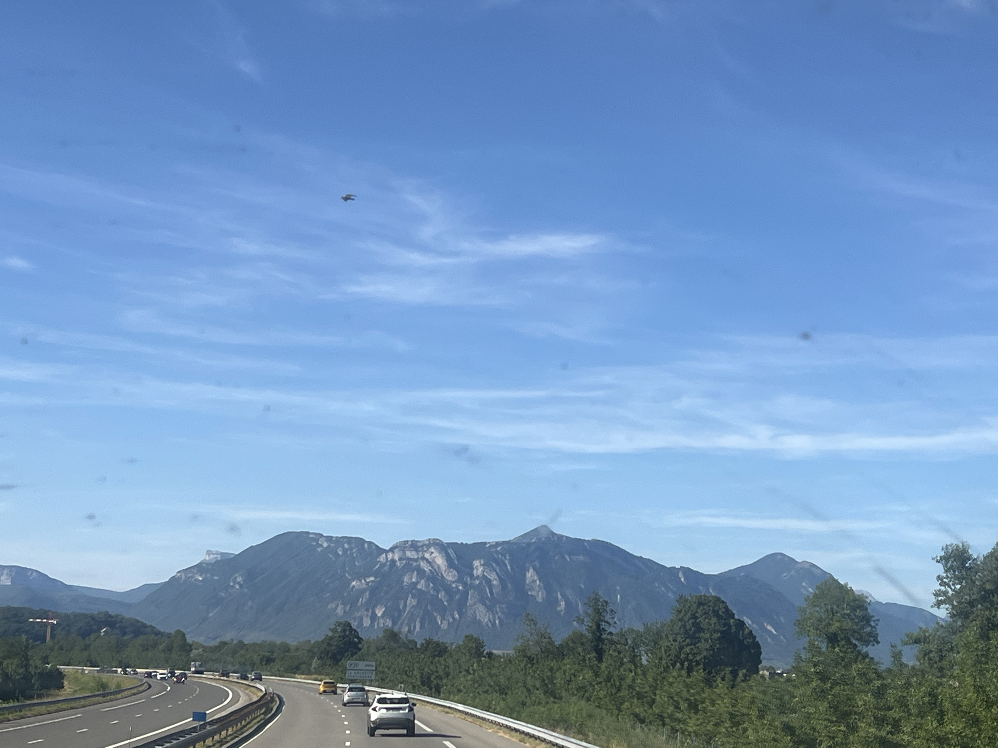 A helicopter is flying in a clear blue sky over a highway in La Buissière, France. The highway has several cars on it. In the background, there are mountains and trees. The scene appears to be captured from a moving vehicle, given the perspective and the slight blur of the landscape. The helicopter is small in the frame, located in the upper left quadrant of the sky.