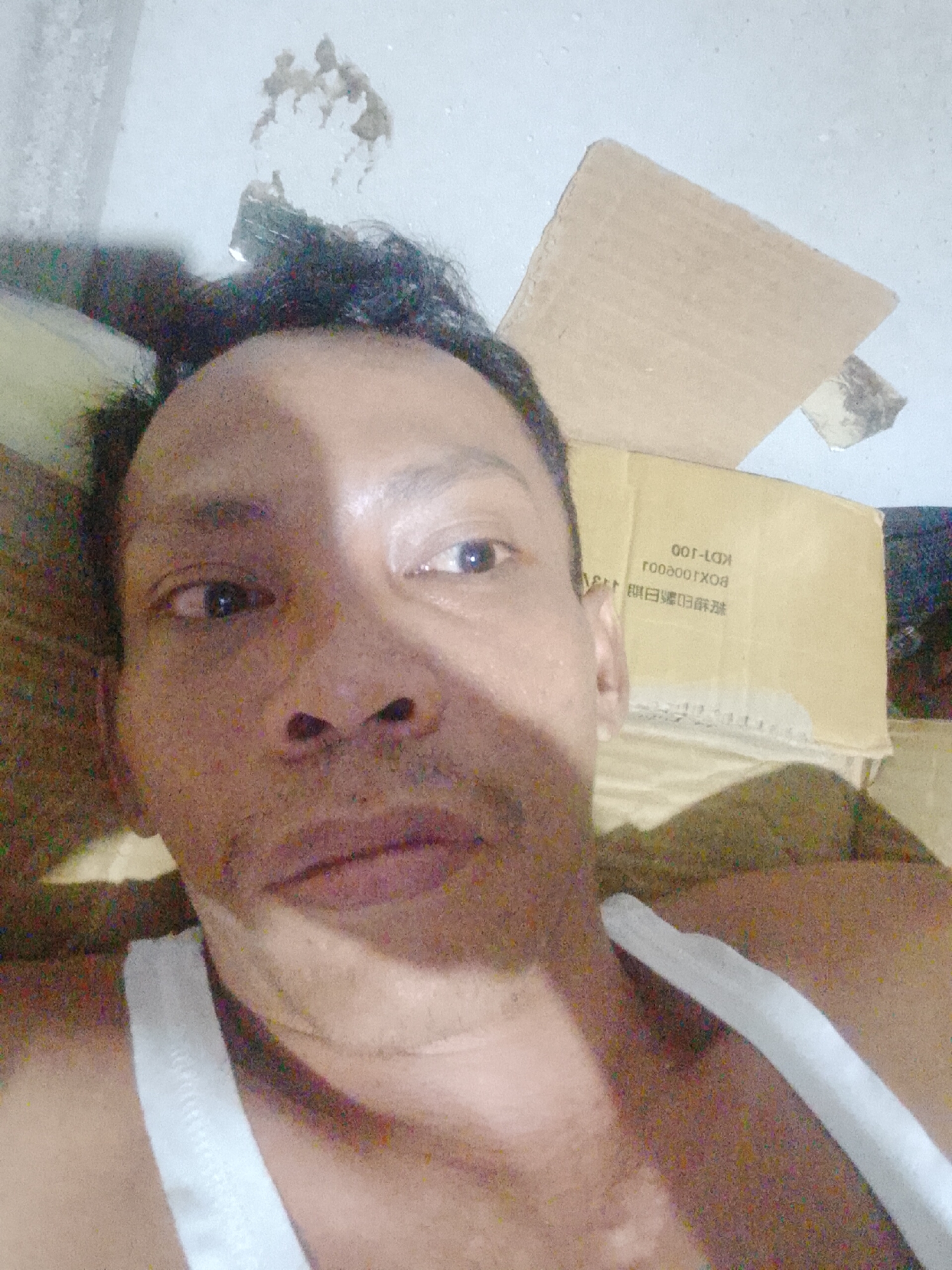 The image is a close-up selfie of a middle-aged man with dark, receding hair and brown skin. He is wearing a white tank top. The background includes cardboard boxes, one of which is partially visible and has text including "KDT-100" and "BOXOOOTXO8". The environment is indoors. The presence of the boxes suggests storage or packing. There is also damage to the wall, indicating a possible state of disrepair. There is no indication of a specific location in Bukit Ketok, Indonesia.