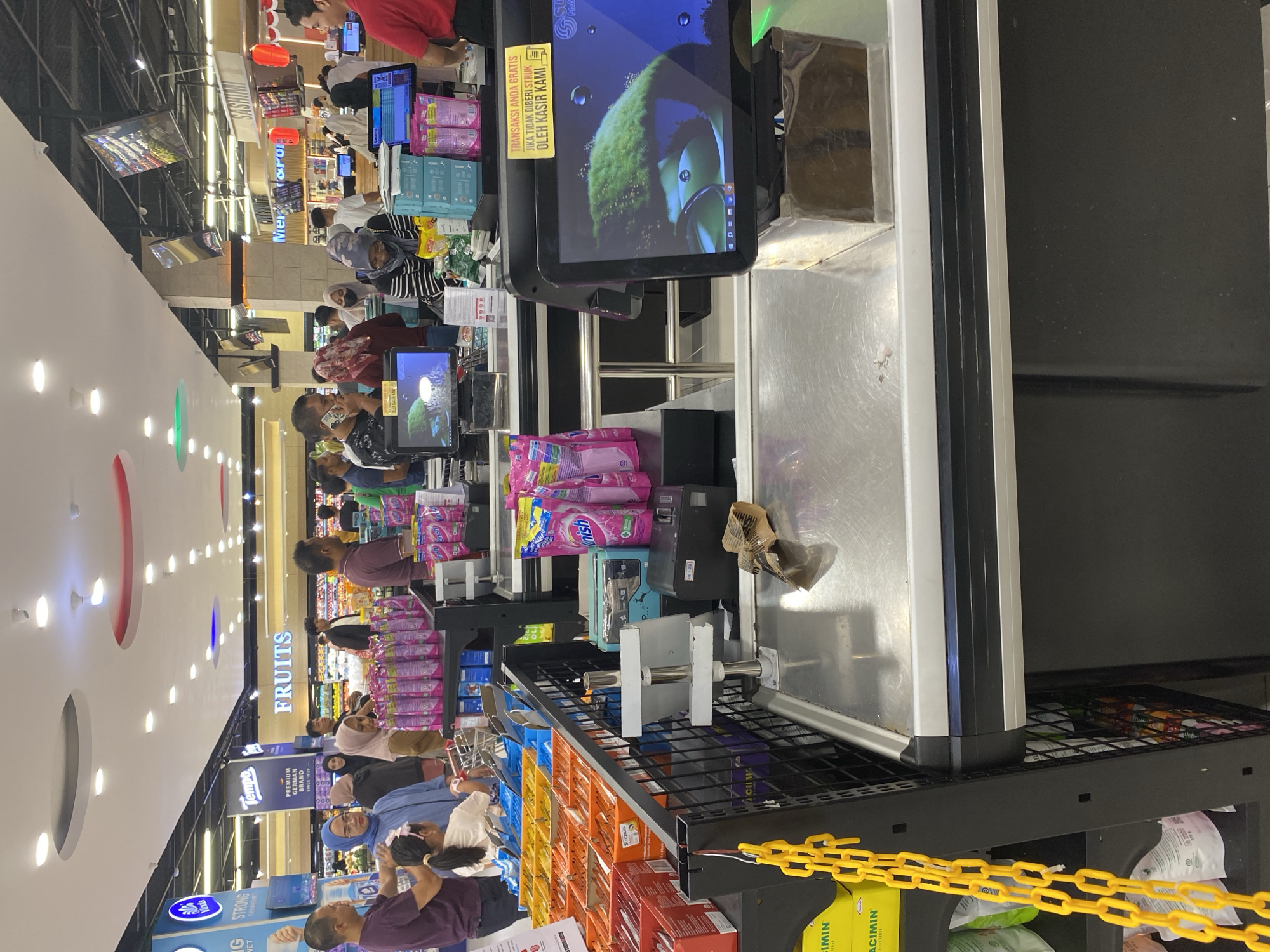 This image depicts a busy checkout area in a supermarket, likely within the city of Banda Aceh, Indonesia. In the foreground, a stainless steel checkout counter is prominent, featuring a cash register system with a touchscreen monitor displaying a green, fuzzy object with water droplets, possibly a screensaver. Adjacent to the monitor are various checkout supplies, including boxes of cleaning products in pink packaging, a printer, and a small white stand with a screen. A yellow chain hangs from the side of the counter.
Behind the counter, several people are engaged in the checkout process. Some are standing in line, while others are interacting with cashiers. The shoppers are diverse, with many wearing headscarves, indicative of the local culture. Among the visible items are groceries and shopping baskets. The supermarket's interior is visible in the background, with brightly lit aisles, signage for "FRUITS" and "SASHIMI," and more shoppers navigating the store. The overall impression is one of a bustling retail environment, with a focus on everyday consumer activity.